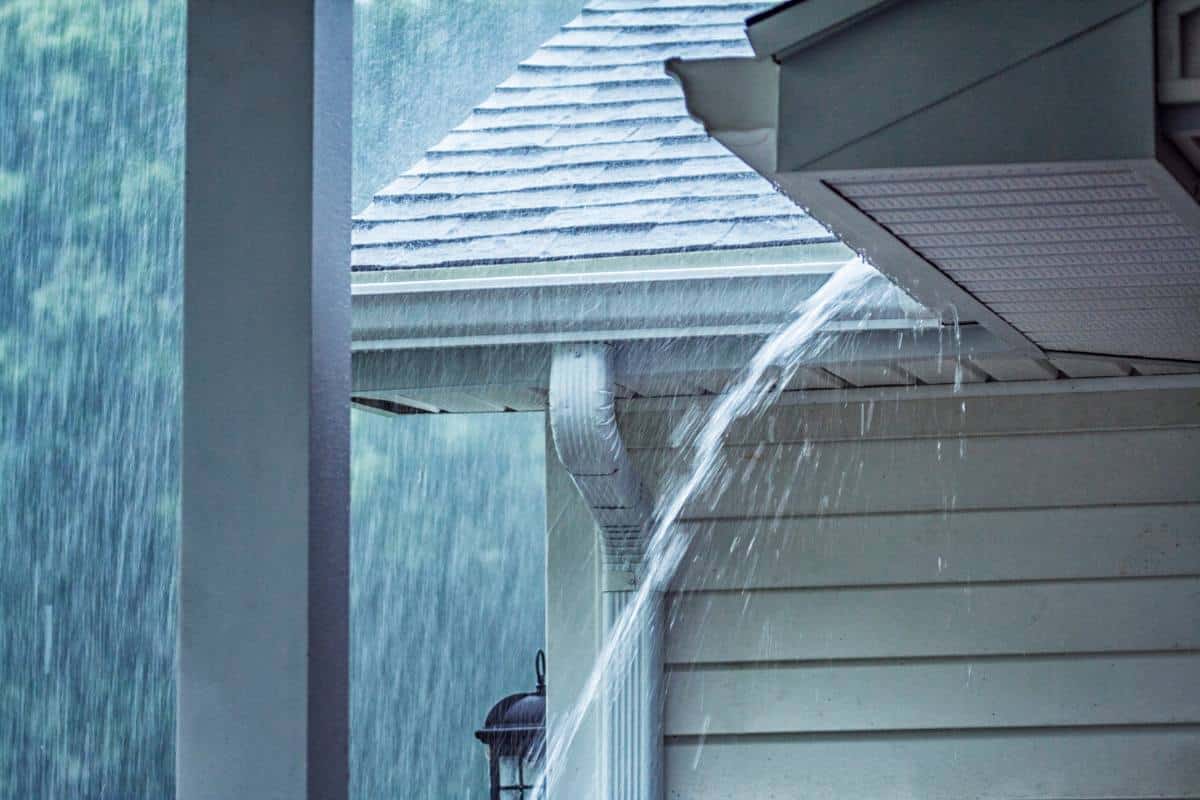 Heavy rainfall splashing against the side of a house, with water flowing from the gutter.