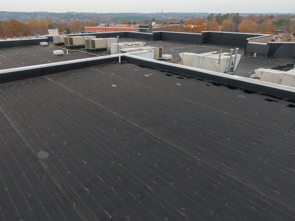 A flat commercial building rooftop with black roofing material, multiple HVAC units, and autumn trees visible in the background under an overcast sky.