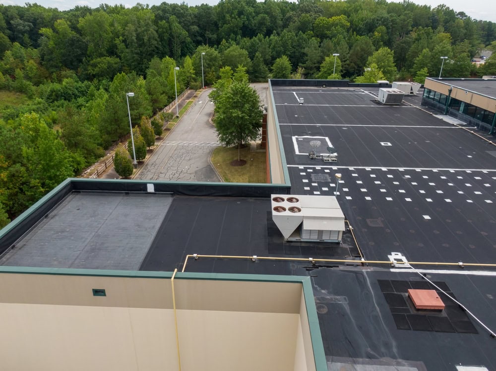 Aerial view of a large, flat-roofed commercial building surrounded by trees and an empty parking lot, with rooftop HVAC units and vents visible.