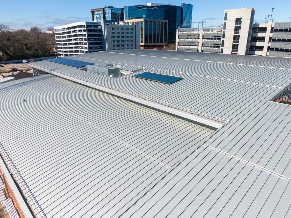 A large, modern metal roof with skylights, surrounded by office buildings, trees, and a multi-level parking structure under a clear blue sky.