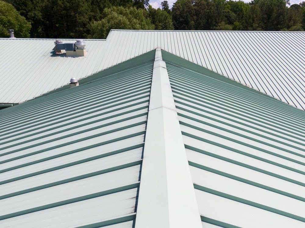 A close-up view of a green metal roof with evenly spaced ridges, showing the roof’s peak and surrounding trees in the background under a clear sky.