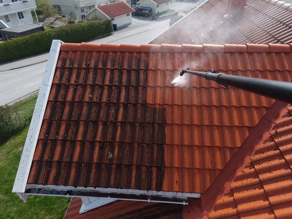 A person is using a pressure washer to clean a tiled roof, removing dark dirt and grime from the tiles and revealing their original bright orange color. Houses and a street are visible in the background.