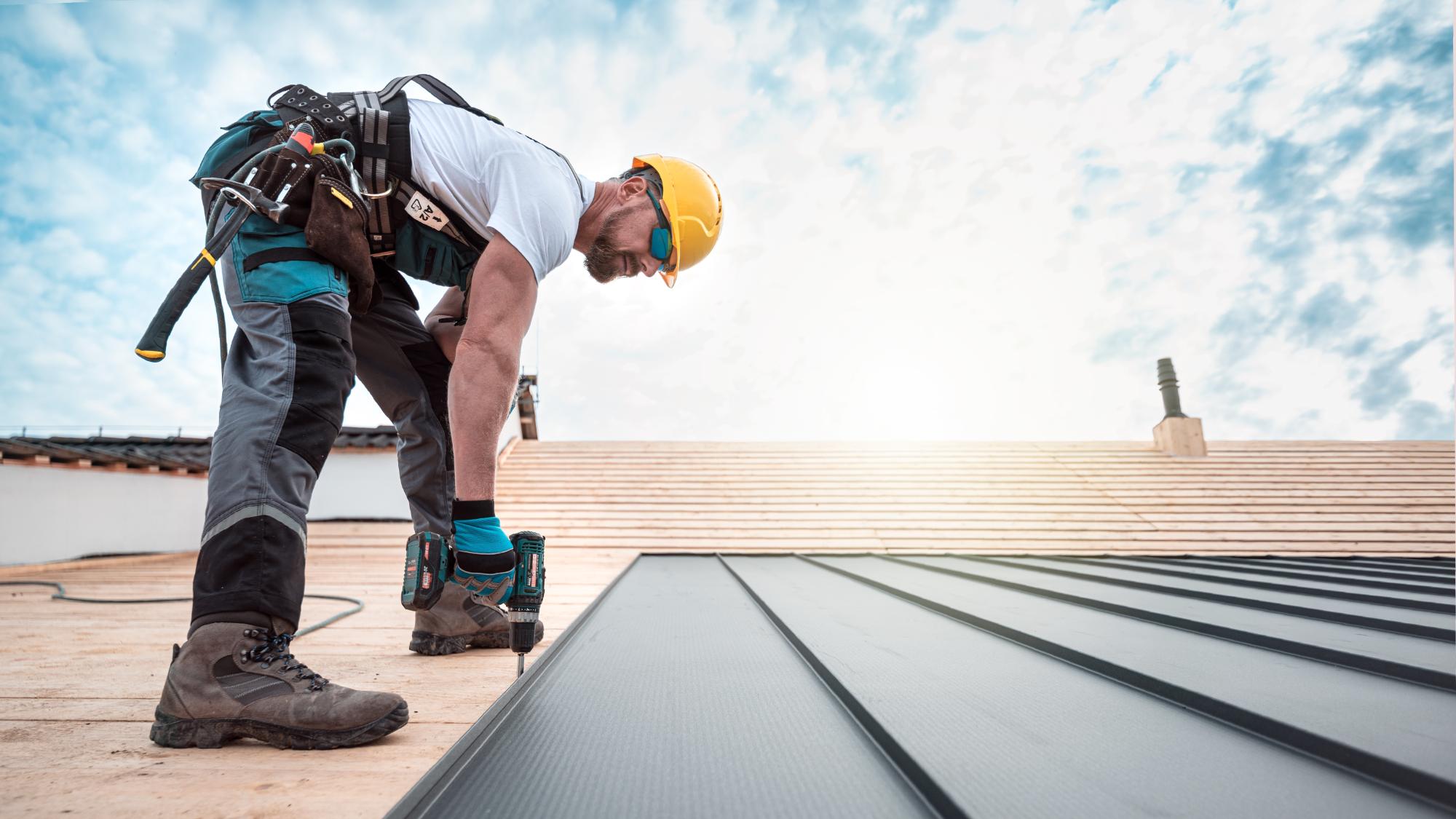 A construction worker wearing a yellow hard hat and safety gear uses a power tool to install metal roofing panels on a wooden roof under a bright, partly cloudy sky.
