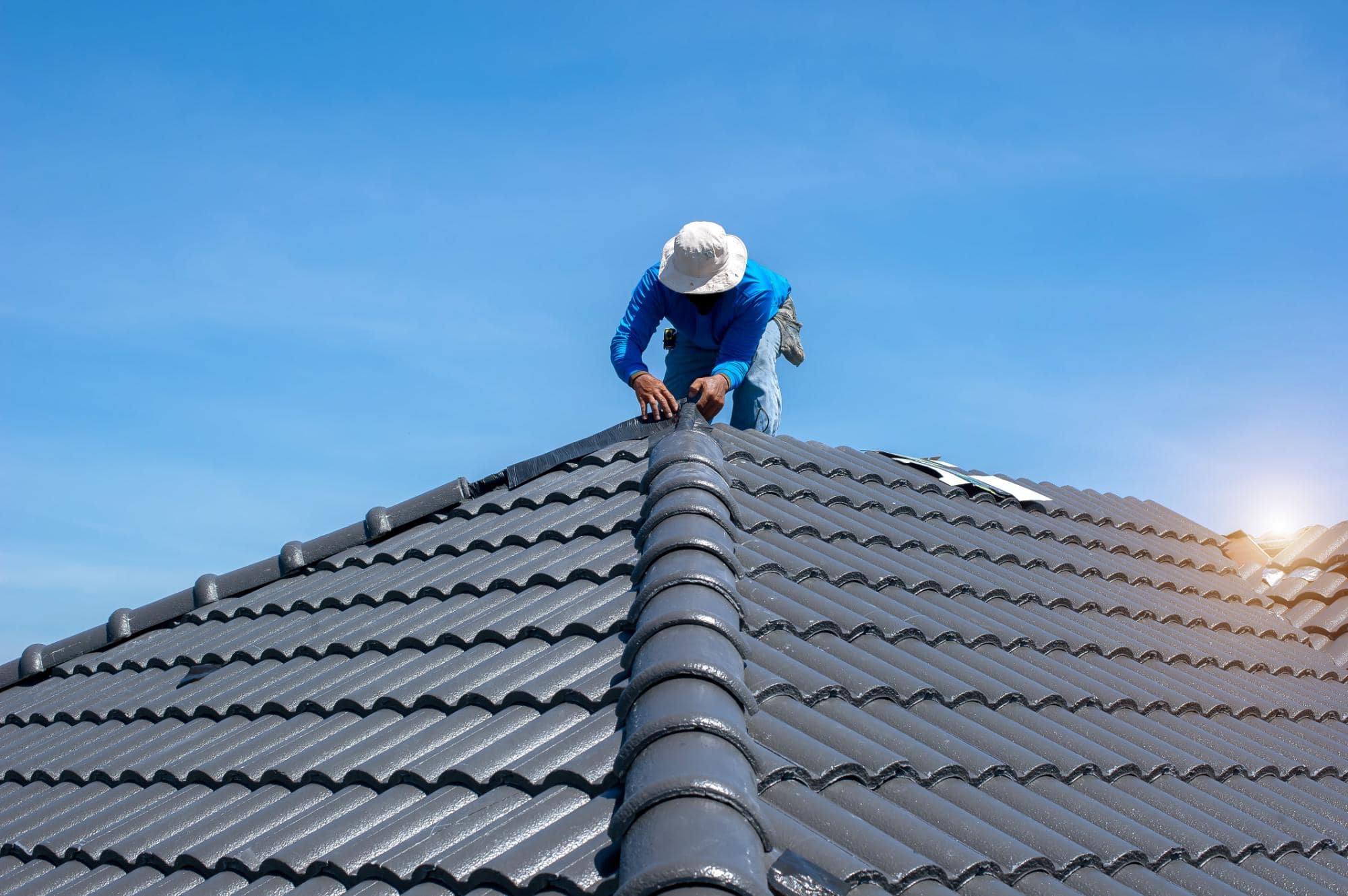 A worker wearing a hat and blue clothing installs tiles on the peak of a grey tiled roof under a clear blue sky, with the sun shining in the background.
