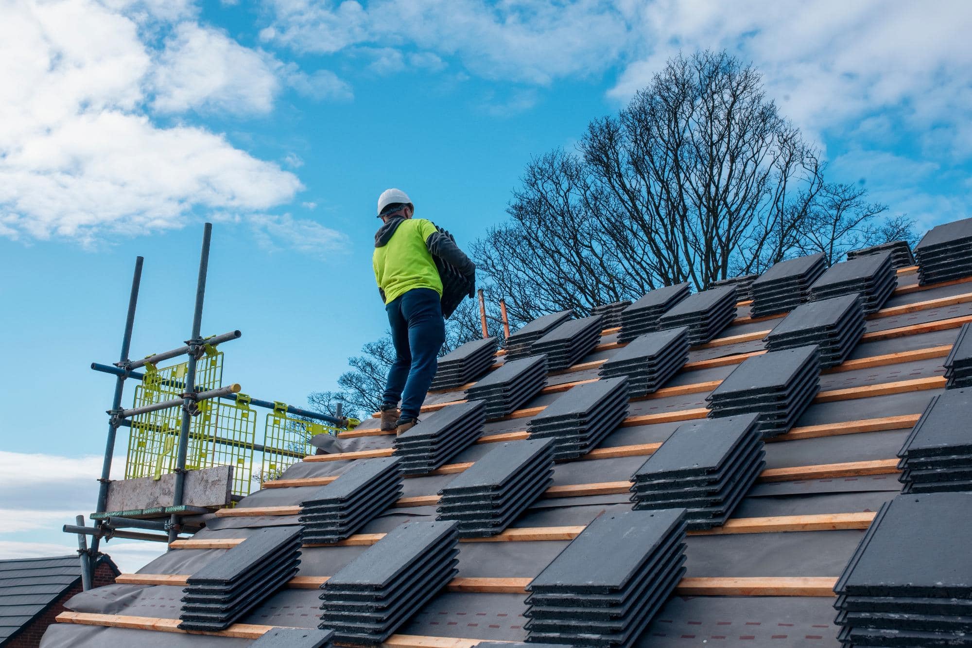 A construction worker in a safety vest and helmet carries black roofing tiles up a partially tiled sloped roof, with scaffolding and leafless trees visible in the background under a blue sky.