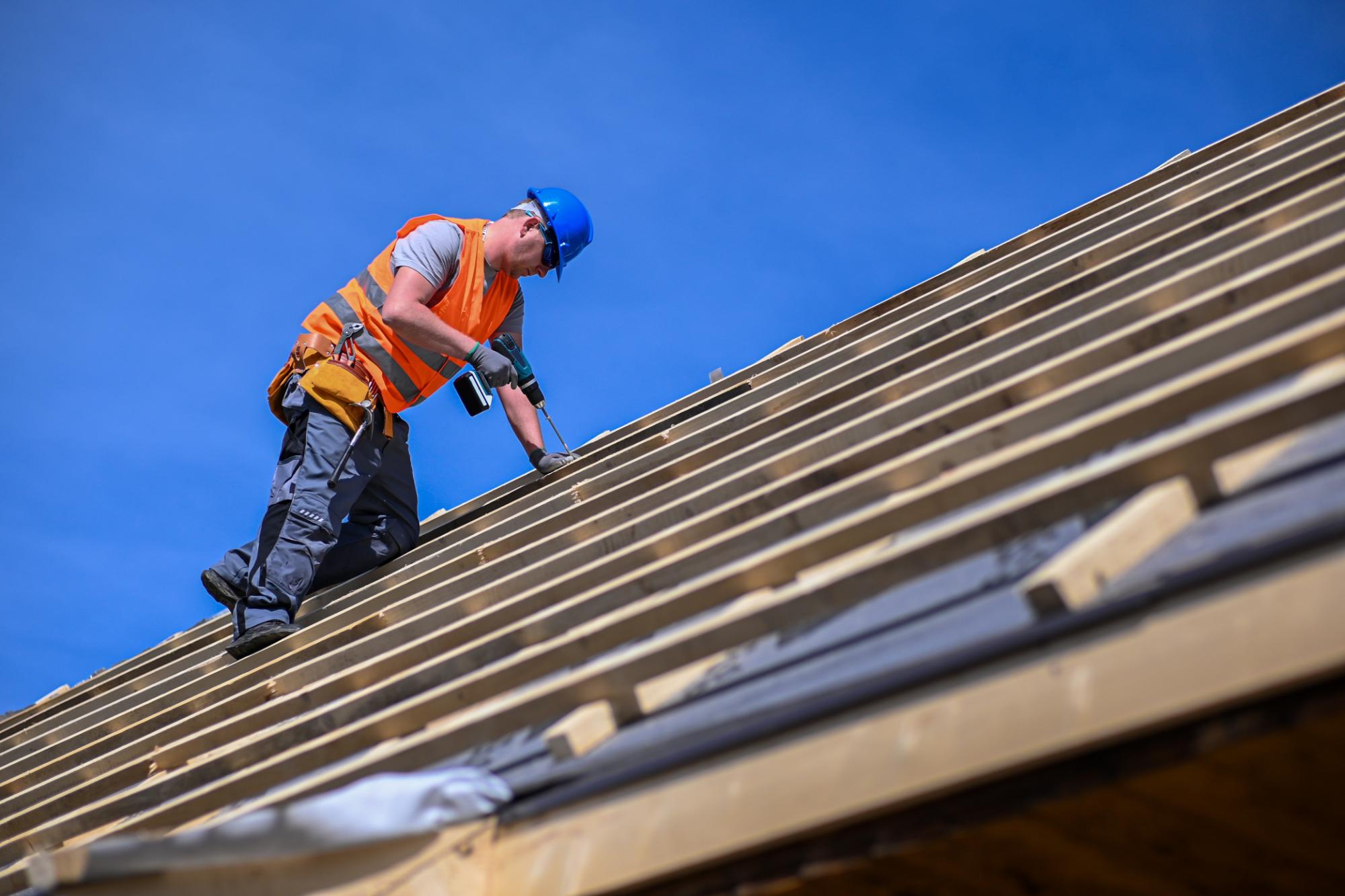 A construction worker in a blue hard hat and orange safety vest uses a tool while working on the wooden framework of a sloped roof under a clear blue sky.