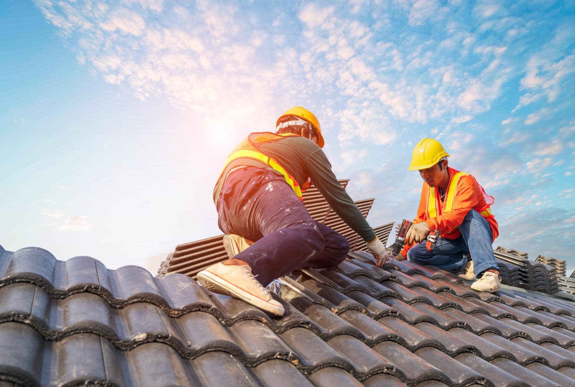 Two construction workers wearing yellow helmets and orange safety jackets install dark roof tiles on a sloped roof under a bright blue sky with sunlight in the background.