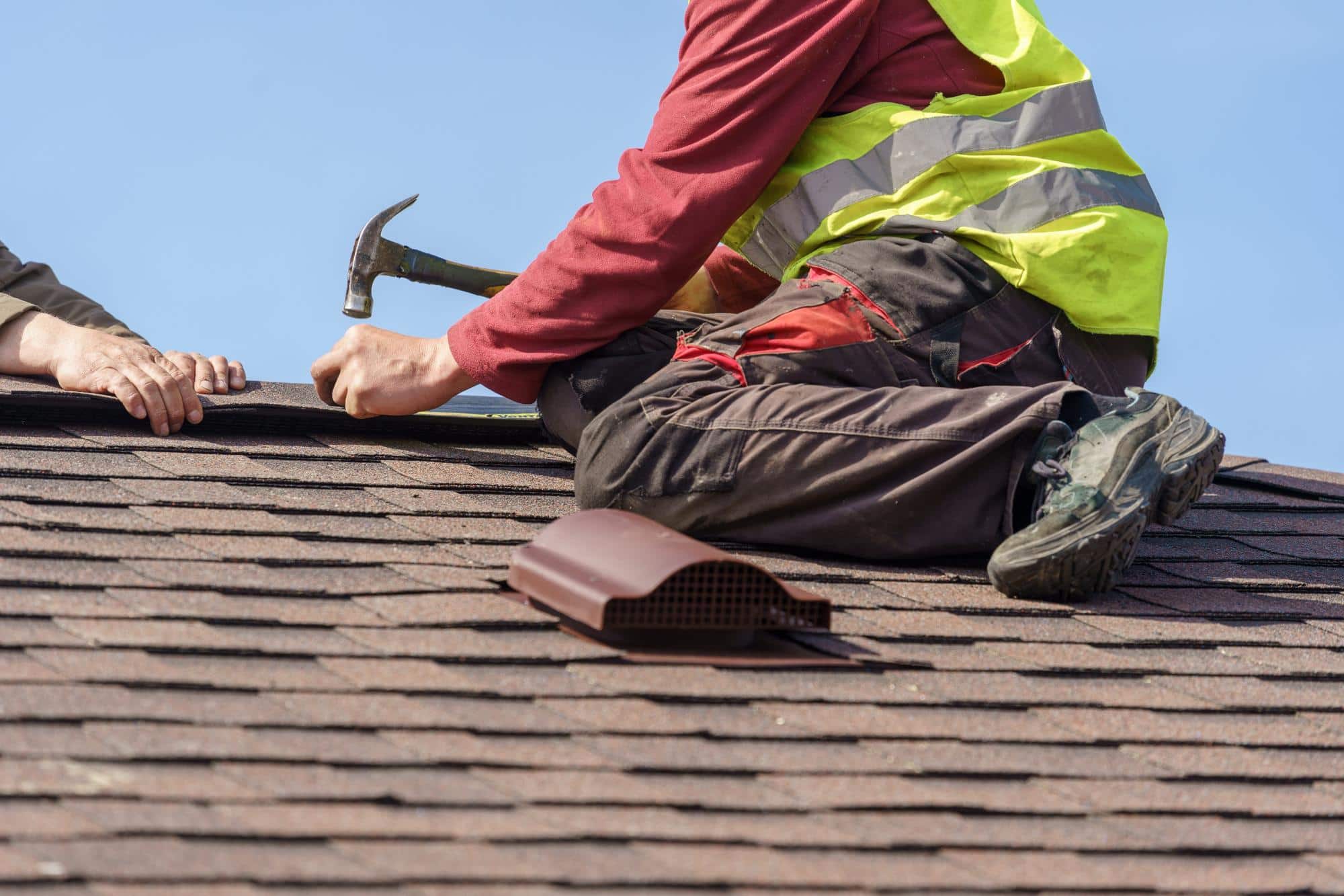 A worker wearing a yellow safety vest and red shirt uses a hammer while installing shingles on a roof. Another person's hand assists nearby. A roof vent rests on the shingles in front of them.