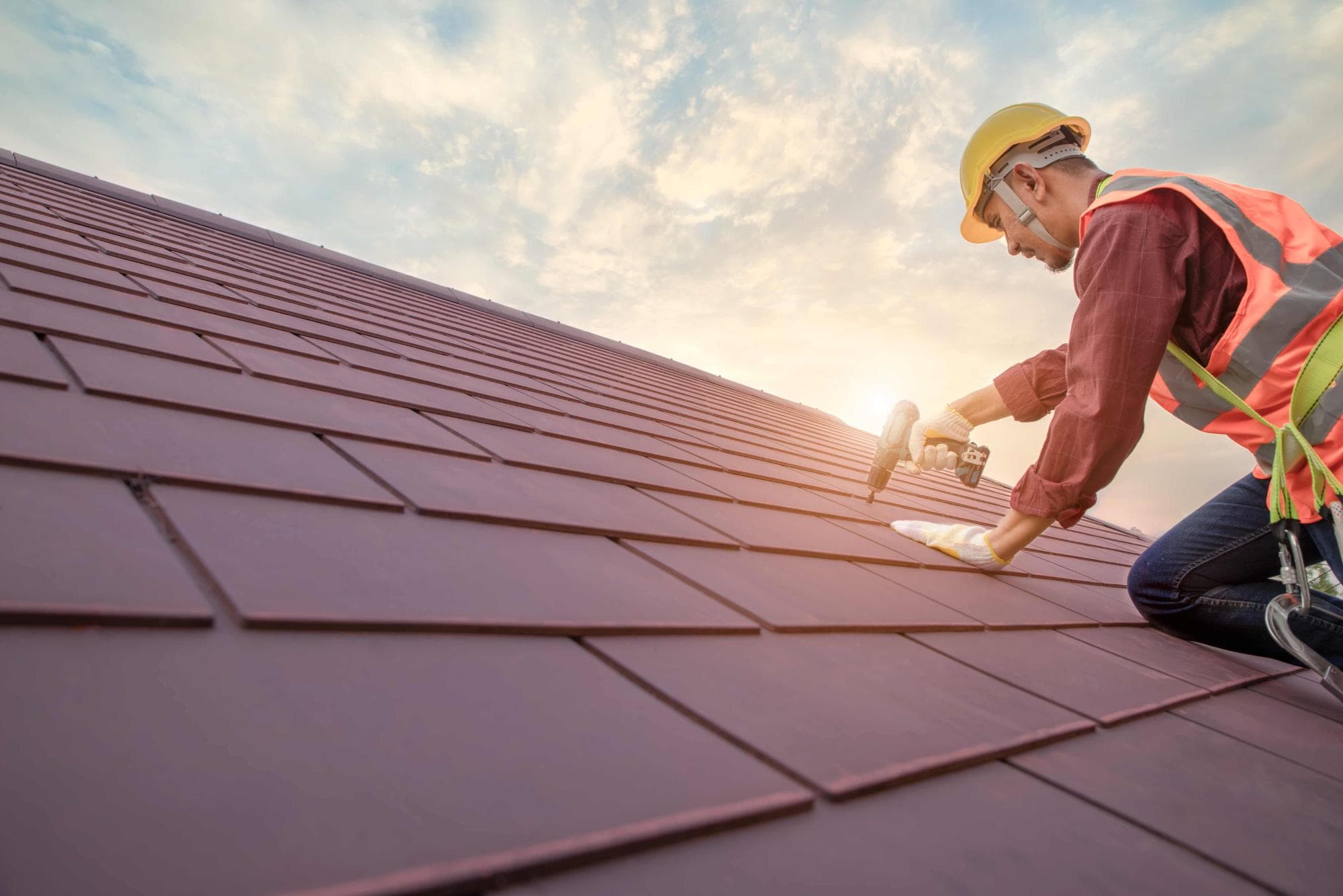 A construction worker wearing a safety helmet and vest uses a power drill while installing or repairing red roof shingles on a sloped roof, with the sun and cloudy sky in the background.