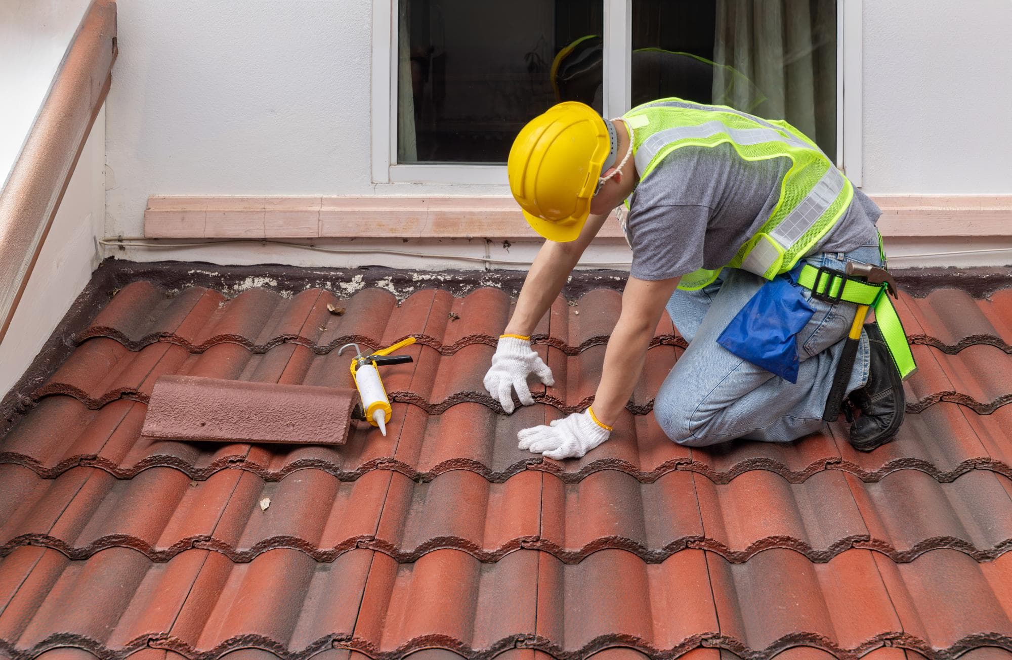 A construction worker in a yellow hard hat and safety vest repairs red roof tiles with tools and sealant next to a window.