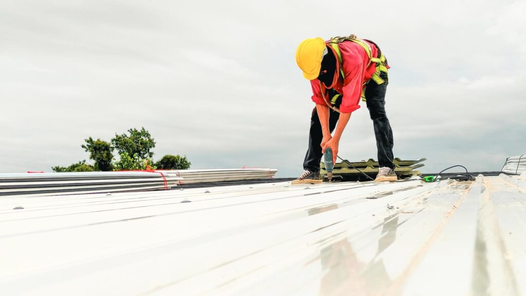 A construction worker wearing a yellow helmet and safety harness installs or repairs a metal roof, using a tool while working outdoors under a cloudy sky. Stacks of metal sheets are nearby.