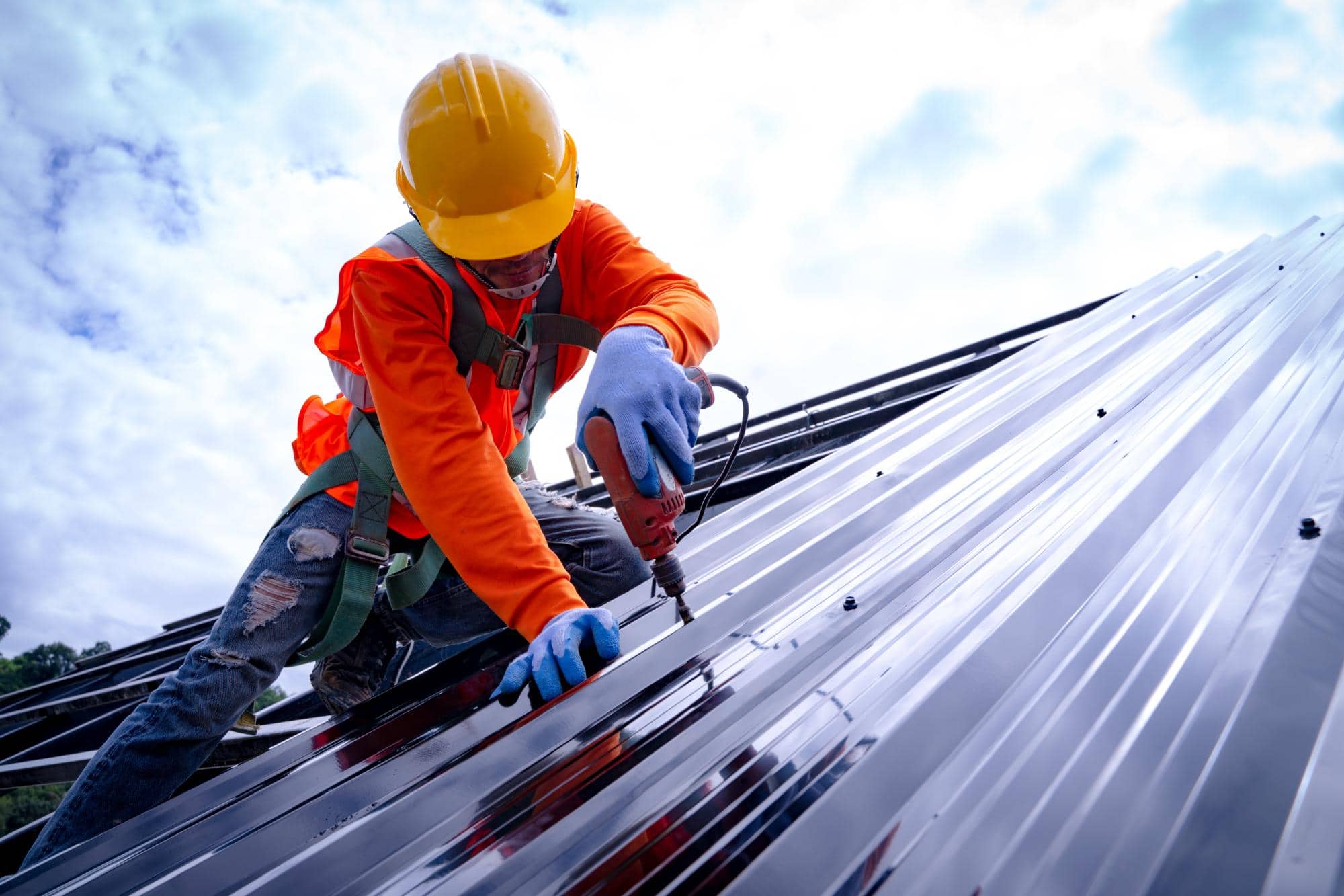 A construction worker in safety gear and a yellow hard hat uses a power drill on a shiny metal roof under a partly cloudy sky.
