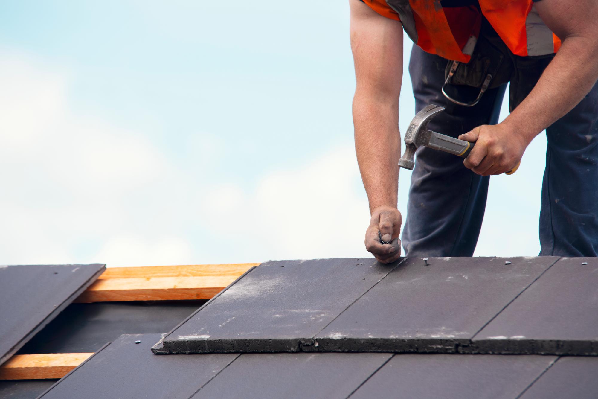 A construction worker in a safety vest uses a hammer to secure dark roof tiles onto a wooden framework under a bright sky. Only the worker’s upper body and arms are visible.