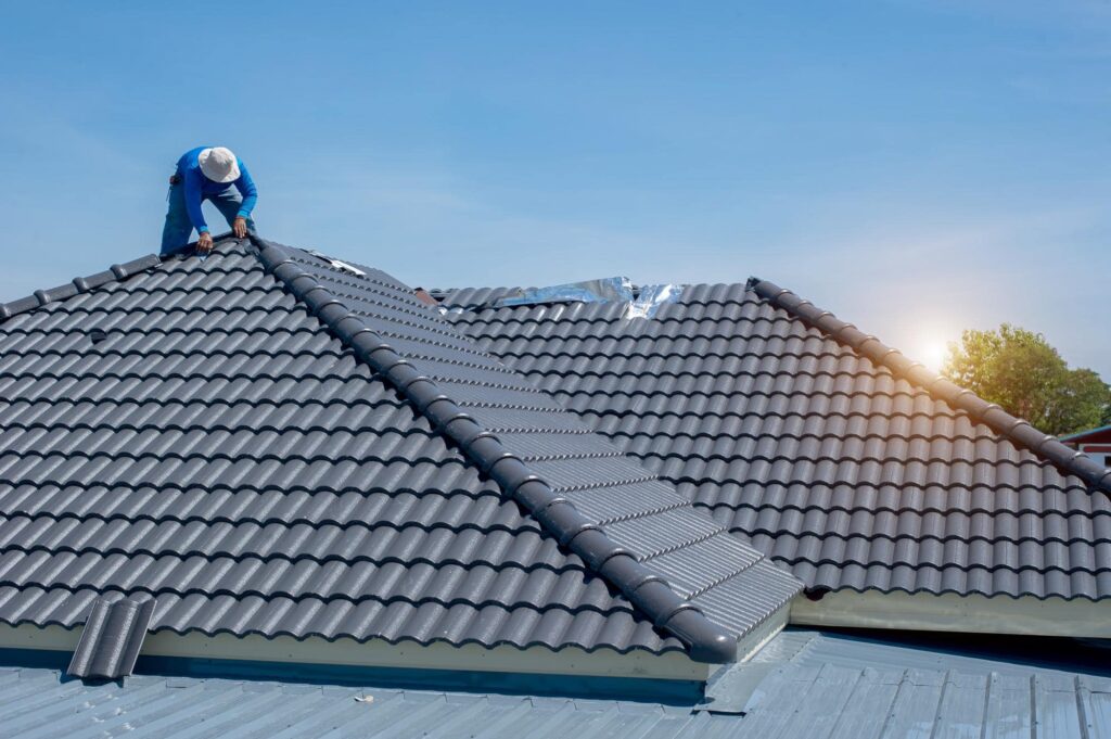 A worker wearing a hat installs dark gray roof tiles on a house under a clear blue sky, with the sun shining in the background.