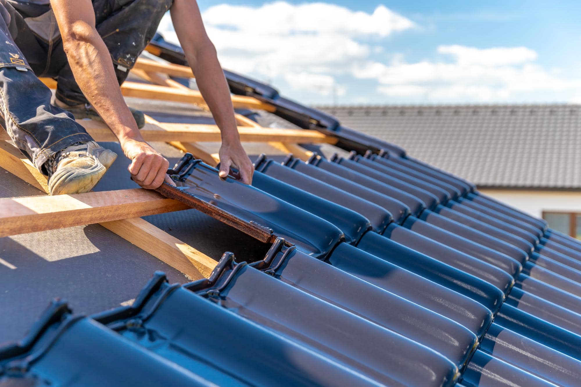A worker installs dark blue roof tiles on a sloped roof frame under a sunny sky, using their hands to position the tiles in neat rows.