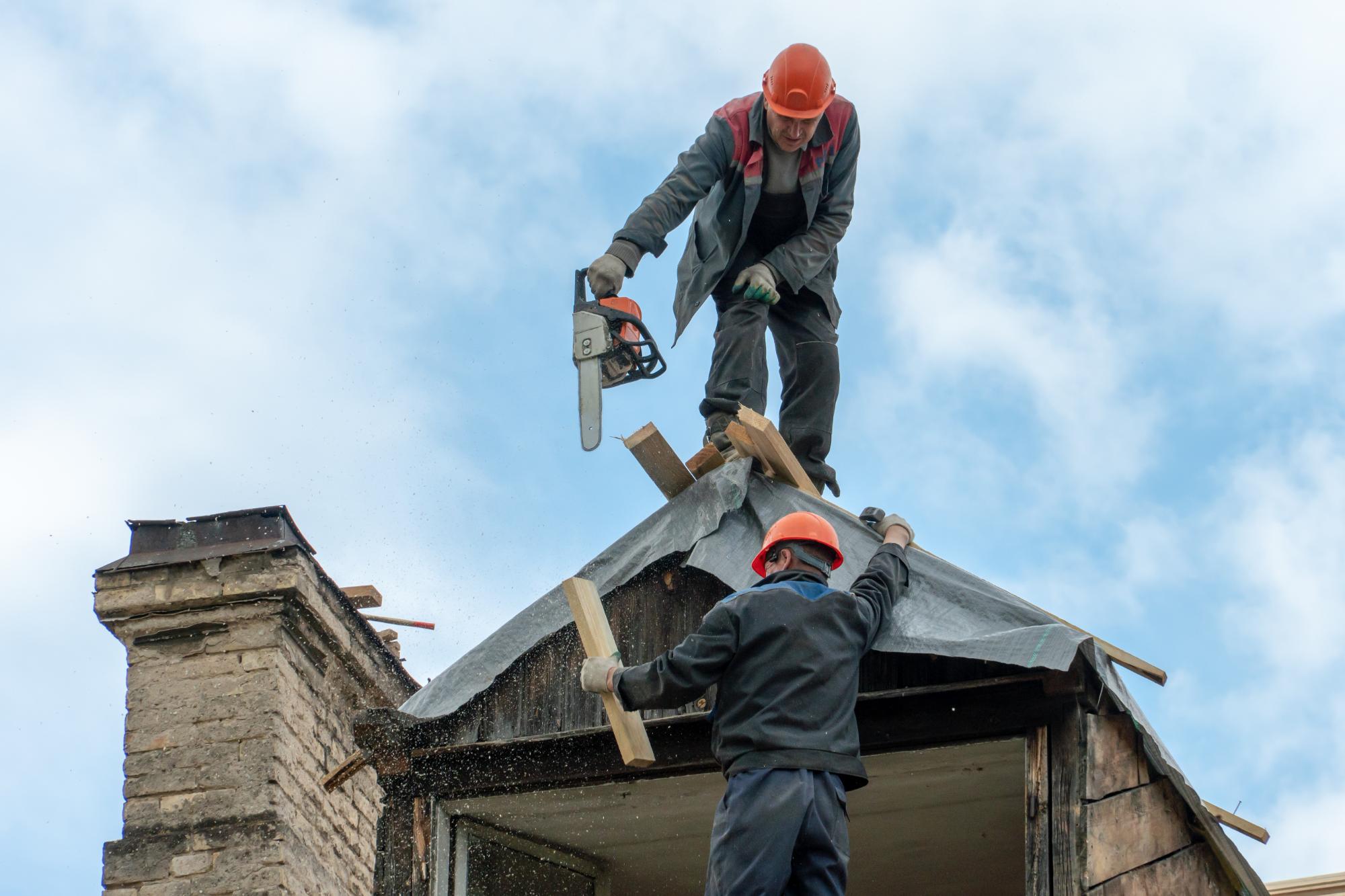 Two construction workers wearing orange helmets are standing on the roof of a building. One worker uses a chainsaw to cut wooden beams, while the other holds the structure steady. The sky is cloudy in the background.
