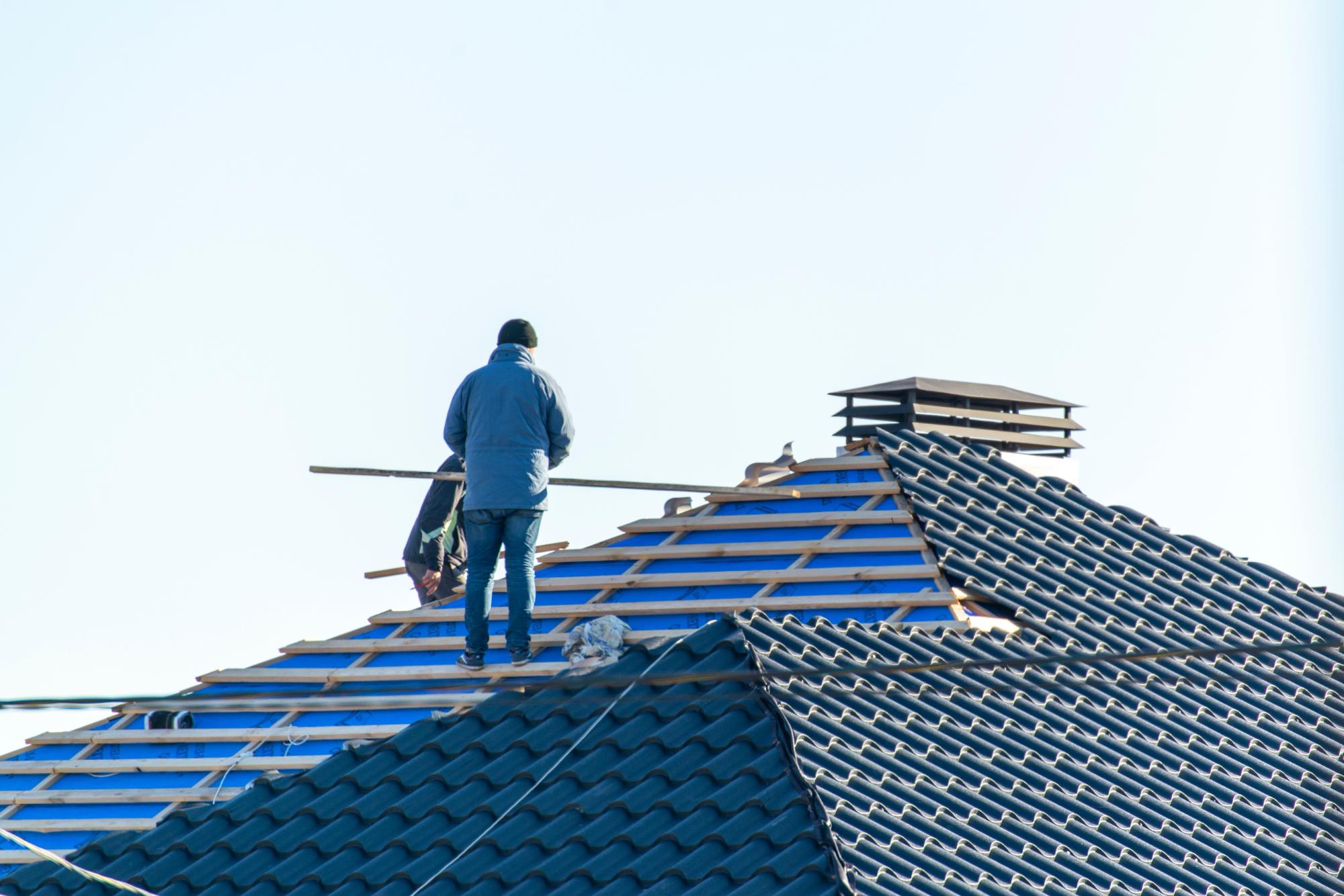 A person in a jacket and hat stands on a partially constructed tiled roof, working with wooden beams on a clear day. The roof has visible construction materials and a chimney.