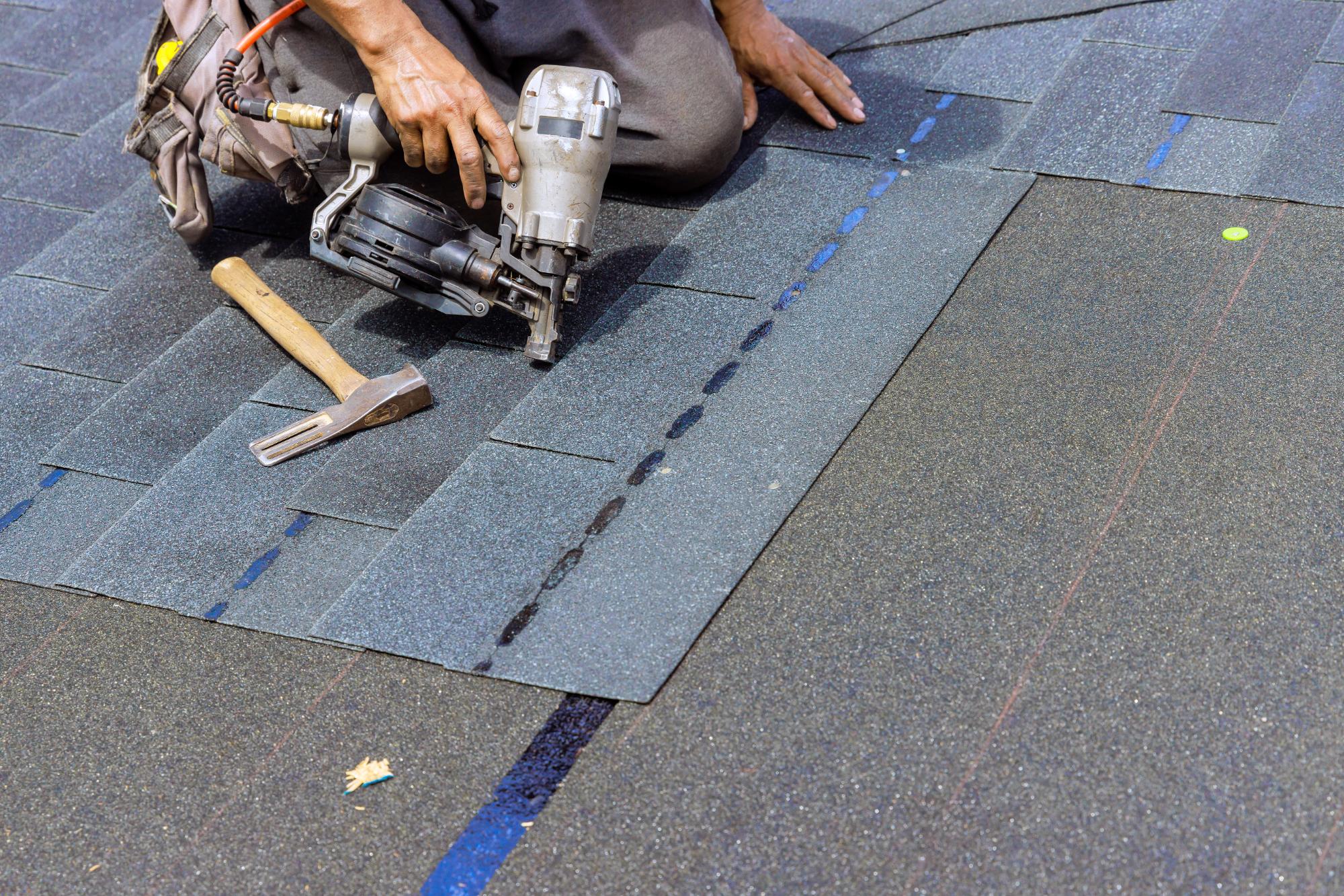 A worker uses a nail gun to install asphalt roof shingles. A hammer lies nearby on the partially finished roof. Only the worker’s hands and part of their legs are visible.