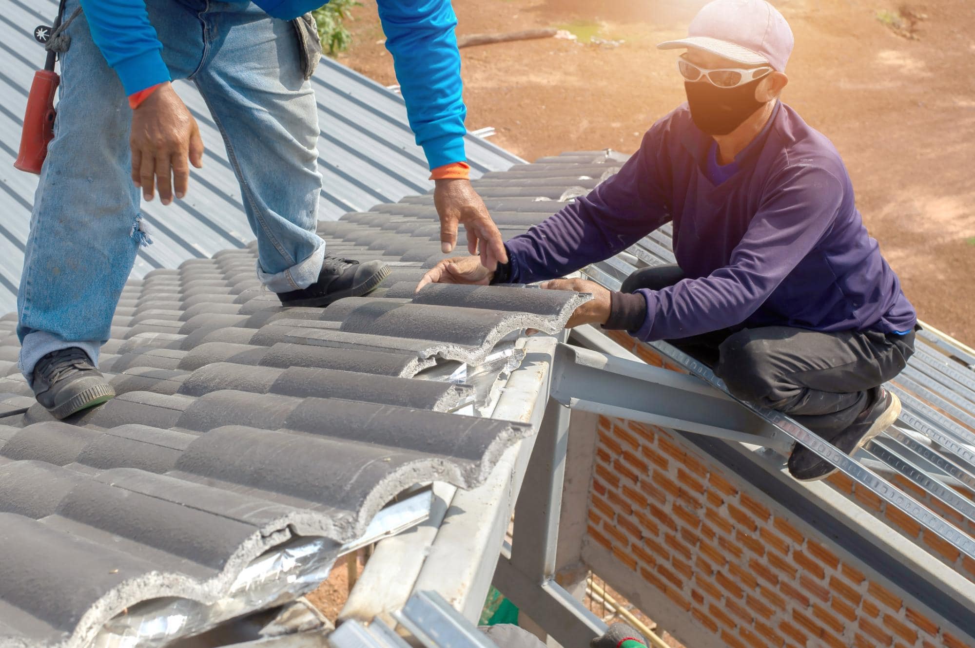 Two workers in protective clothing and masks install gray roof tiles on a house, one kneeling and one standing on the edge of the roof, with sunlight shining on the scene.