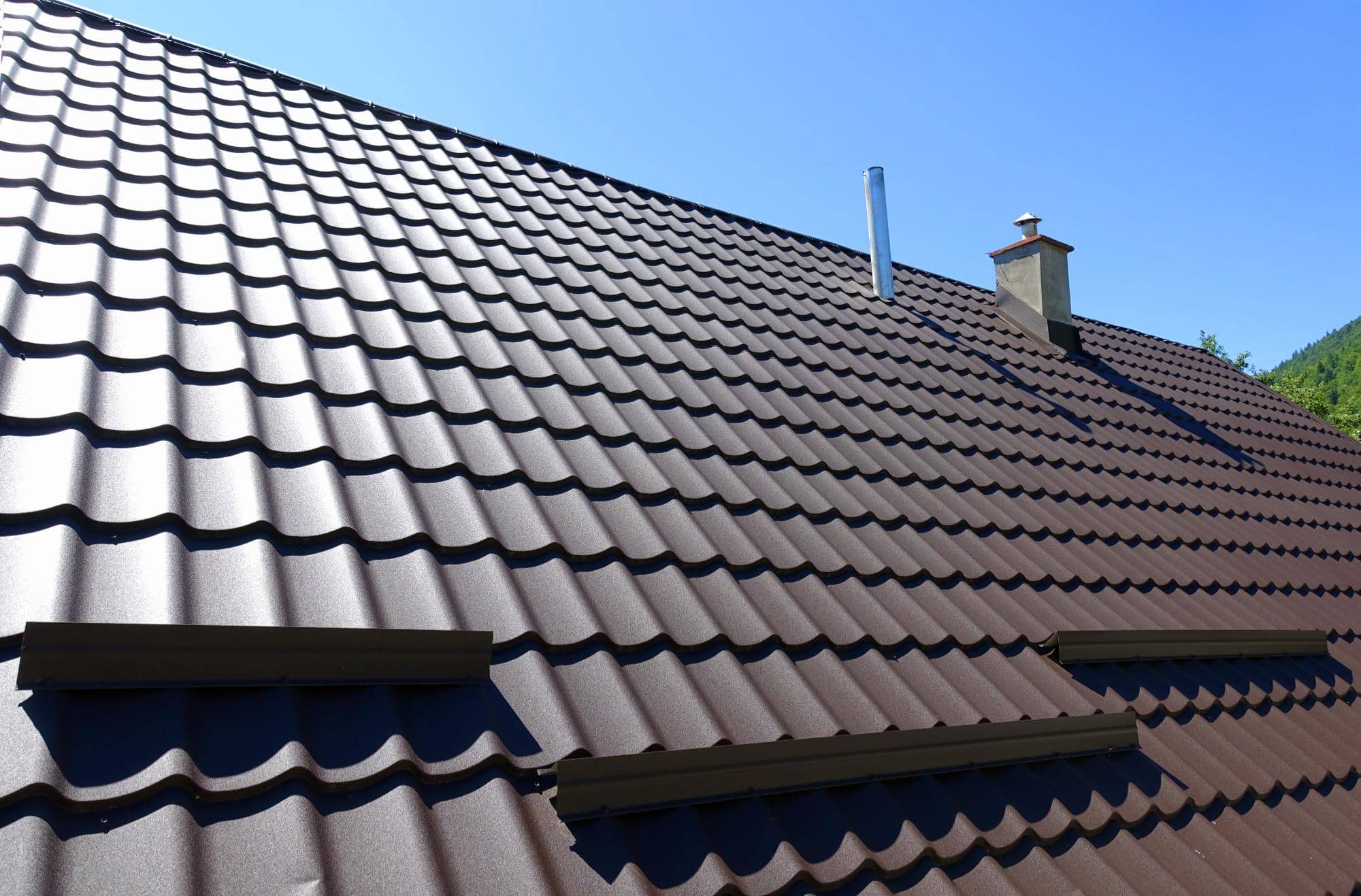 A close-up view of a brown metal tiled roof with a chimney and a pipe, under a clear blue sky. The roof features a wavy design and ventilation openings.