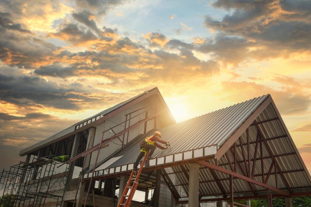 A construction worker on a ladder installs metal roofing on a partially built house at sunset, with dramatic clouds and sunlight in the background.