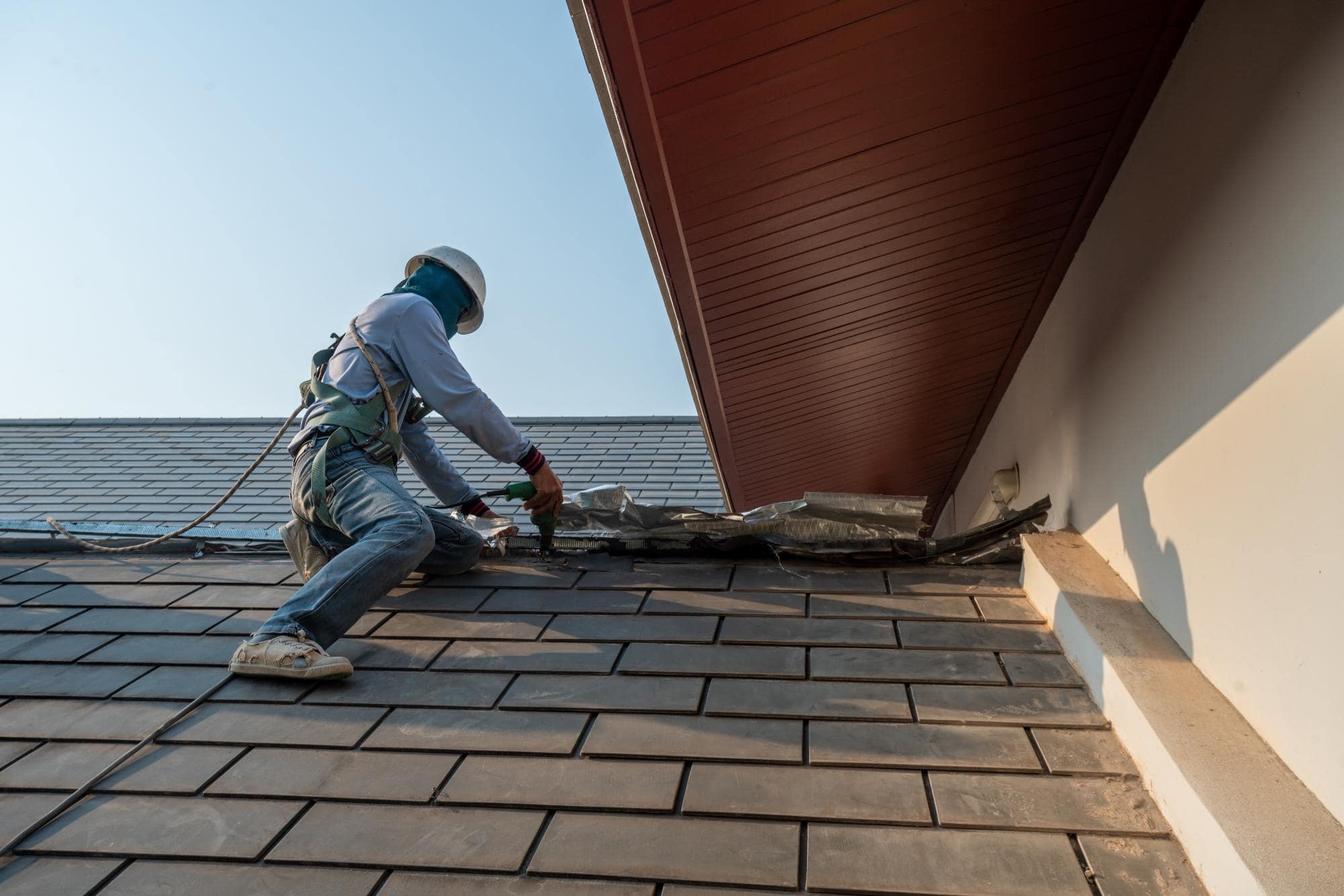 A construction worker wearing safety gear uses tools to install or repair shingles on a house roof, secured with a harness under a clear sky.