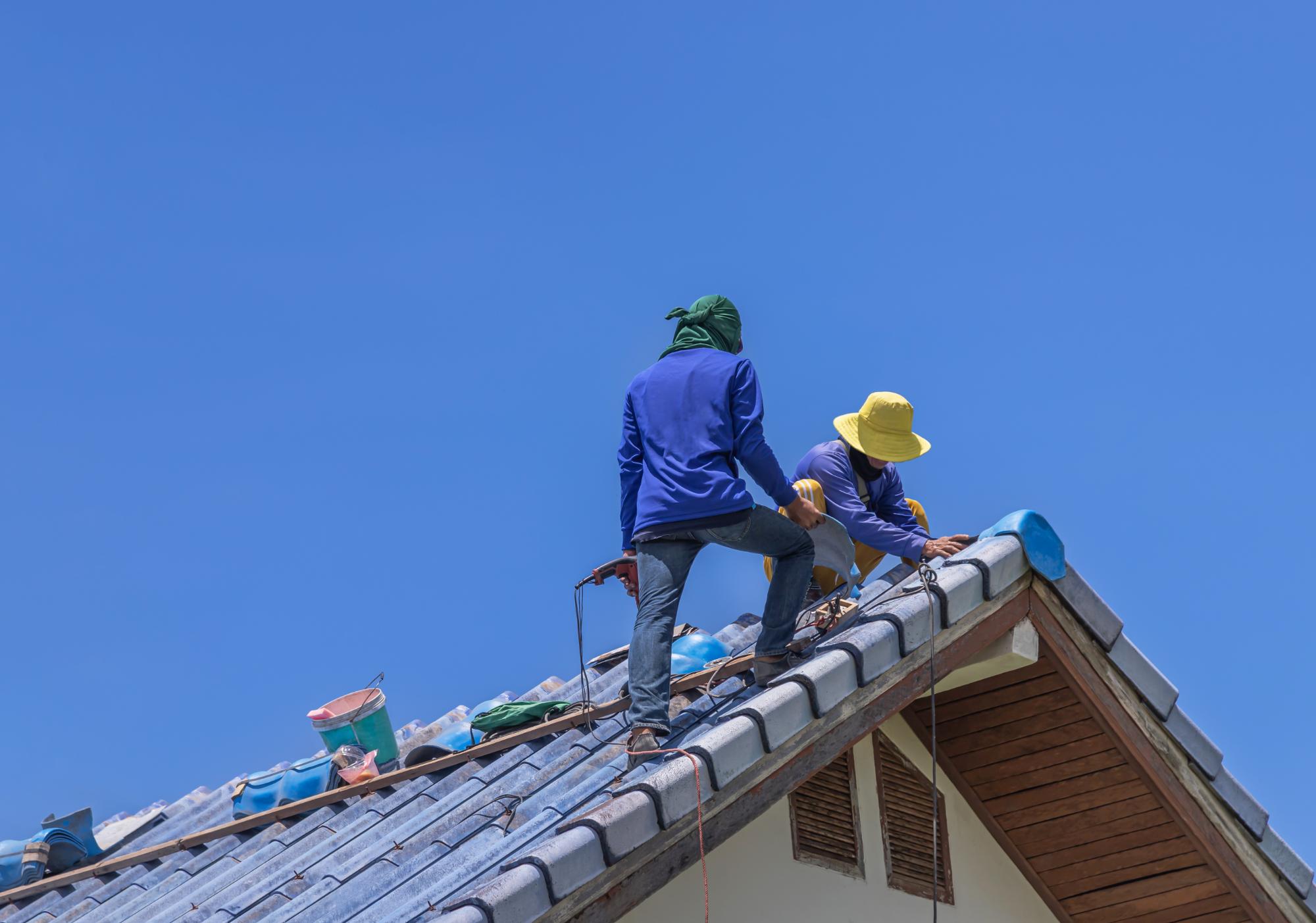 Two workers in hats and protective clothing install or repair blue roof tiles on a house under a clear blue sky. Tools and materials are visible on the roof.