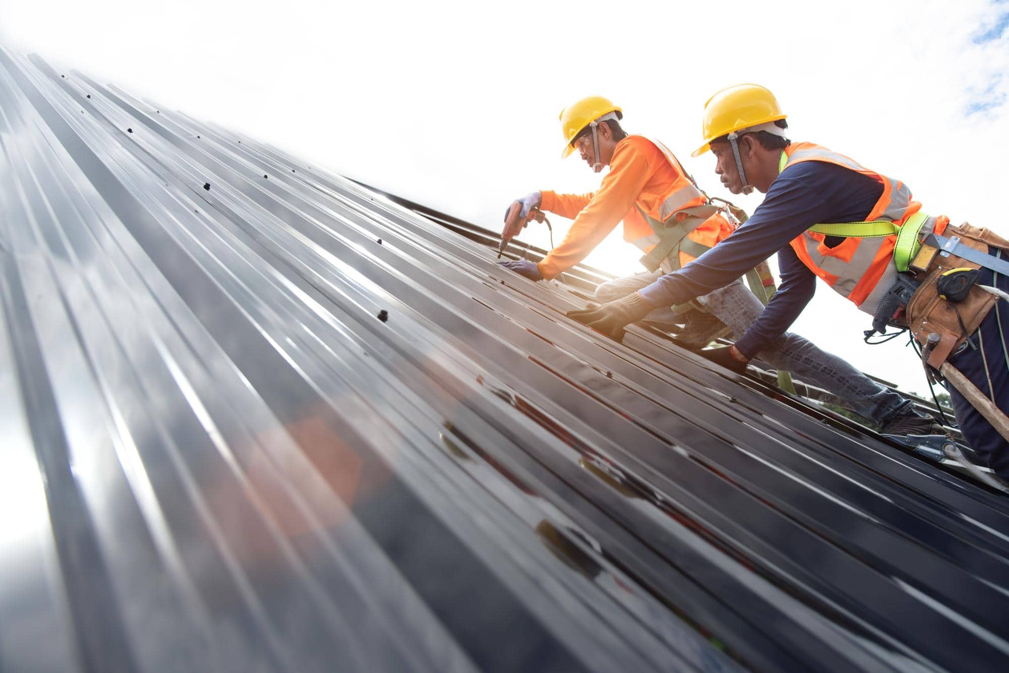 Two construction workers wearing safety gear install metal sheets on a sloped roof under bright daylight. Both are using tools and wearing helmets, gloves, and safety harnesses, working together on the structure.