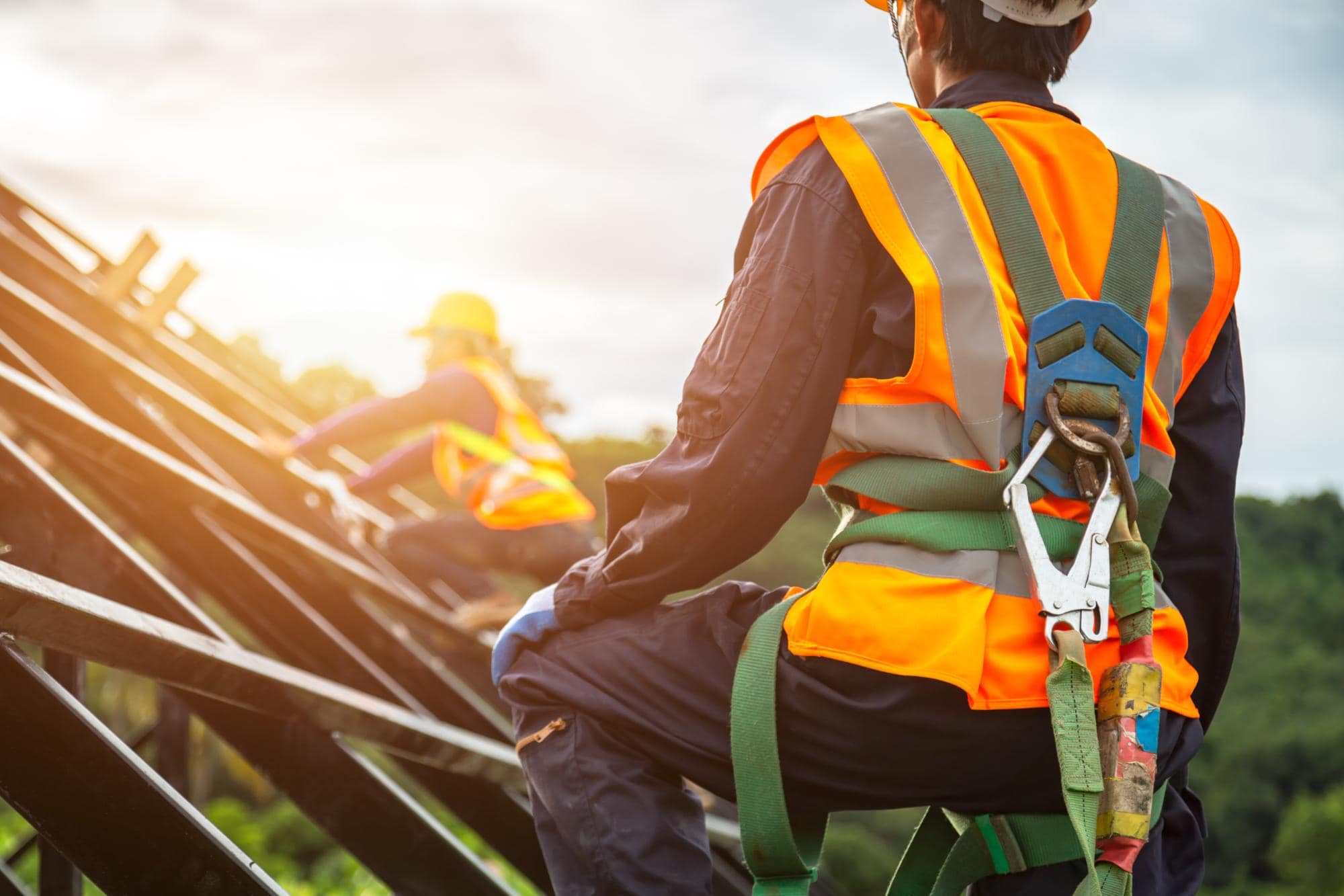 Two construction workers wearing safety harnesses and reflective orange vests work on a metal structure outdoors, with one worker in the foreground and another in the background under bright sunlight.