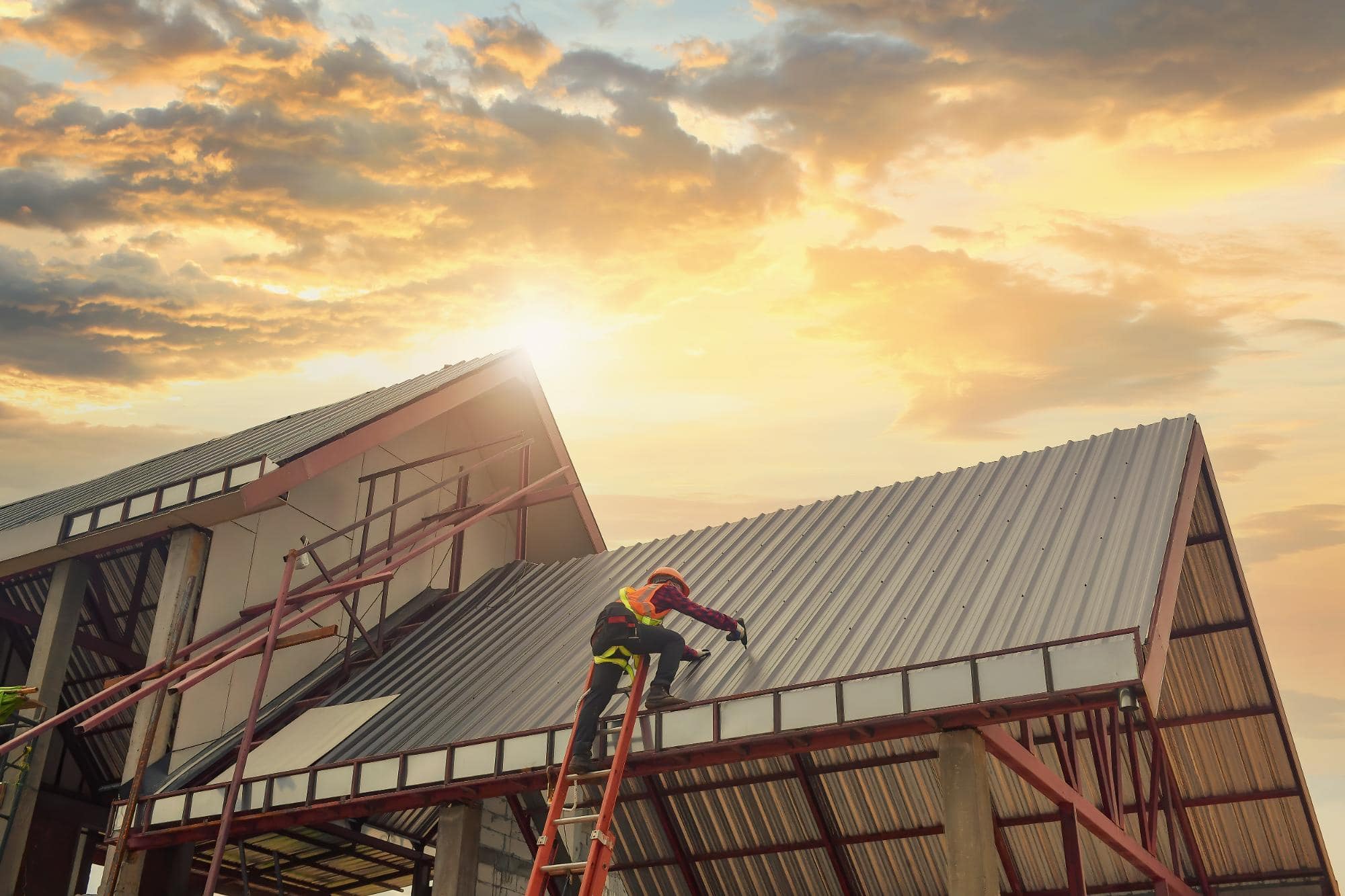 A construction worker on a ladder installs a metal roof panel on a building at sunset, with another partially built structure and a cloudy sky in the background.