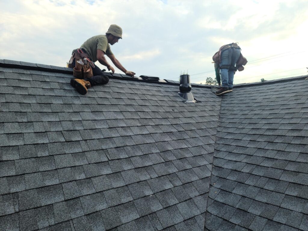Two workers wearing tool belts are installing or repairing shingles on a sloped roof under a cloudy sky. One worker is sitting, and the other is bending over near a roof vent.