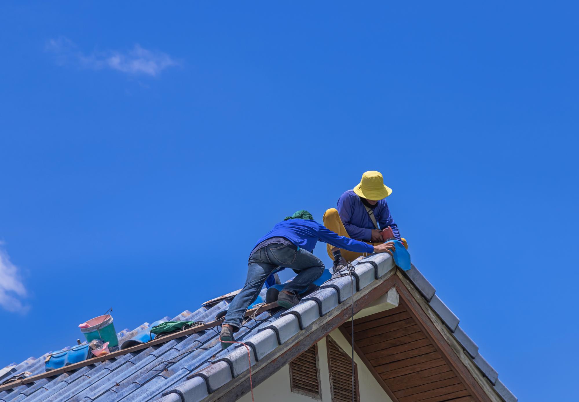 Two workers wearing hats and safety gear are fixing or installing blue roof tiles on a sloped house roof under a clear blue sky. Tools and materials are scattered along the roof.
