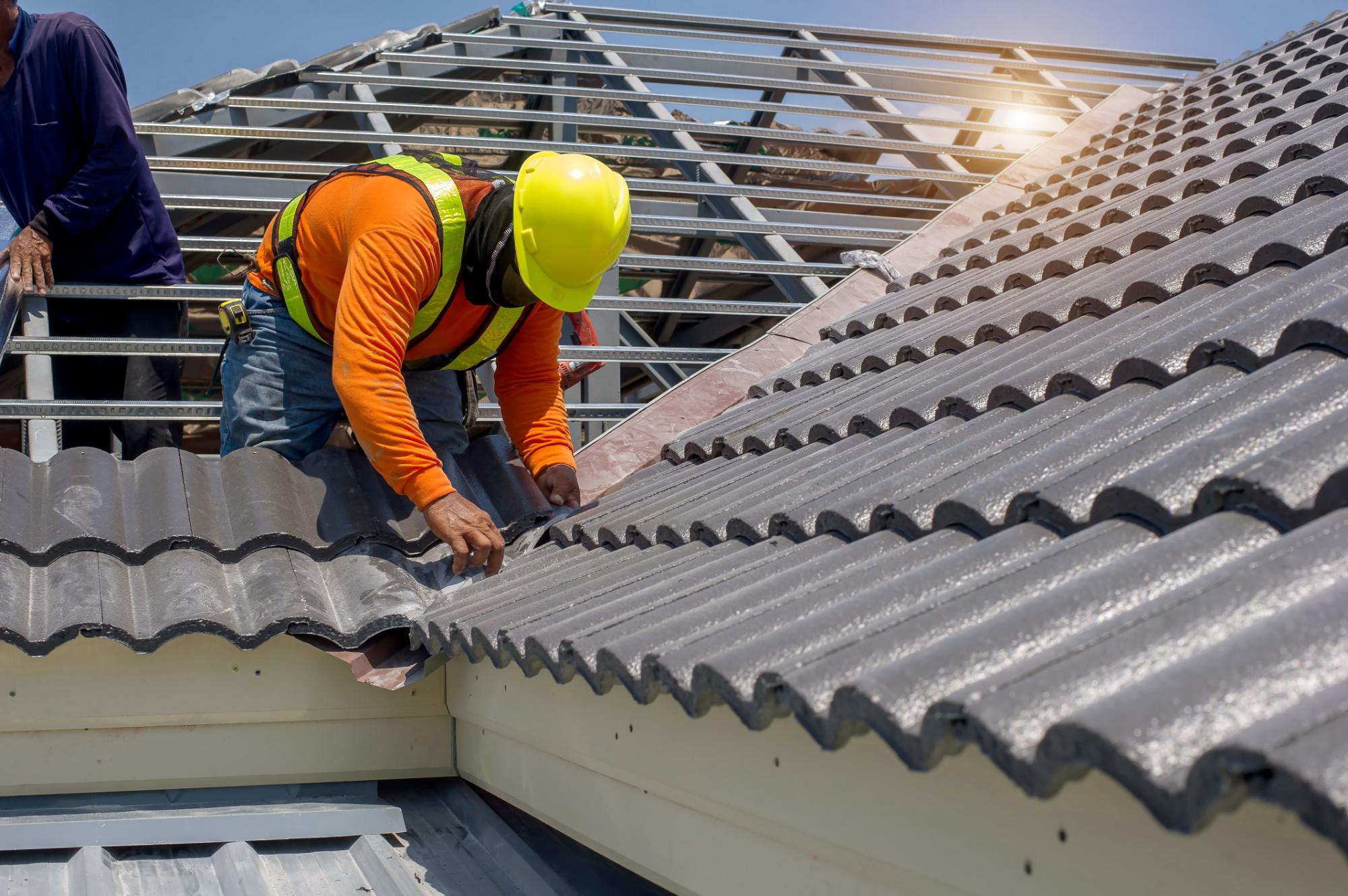 A construction worker in a yellow hard hat and safety vest installs gray roofing sheets on a house roof under a clear sky, with another worker nearby and sunlight shining in the background.