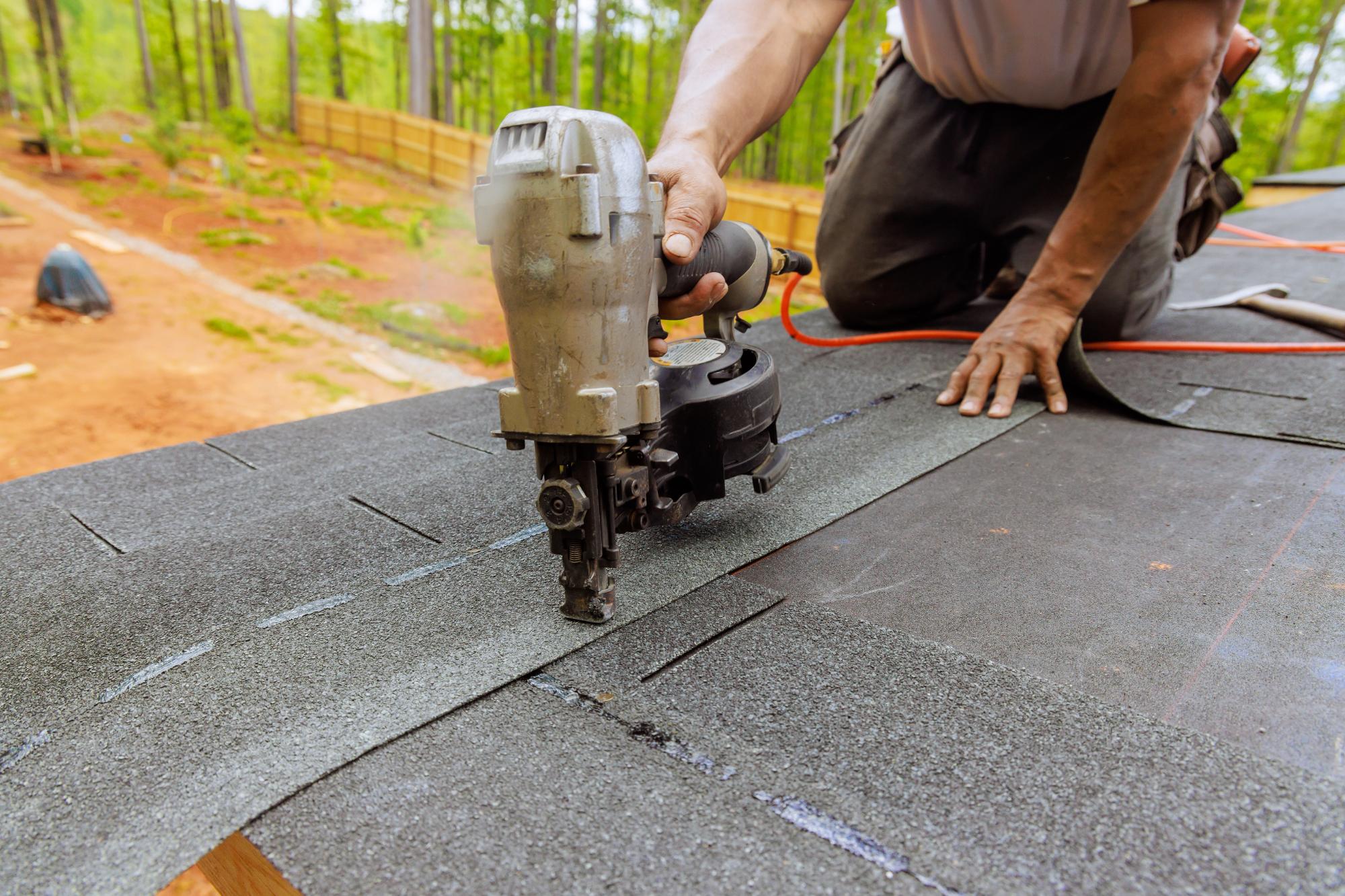 A person uses a nail gun to secure roofing felt on a roof, with one hand guiding the tool and the other pressing down on the material; trees and a fenced yard are visible in the background.