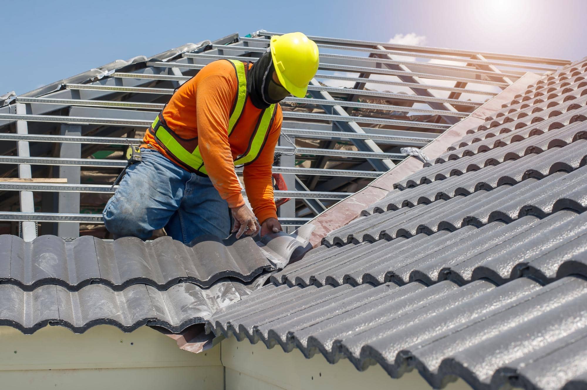 A construction worker in a yellow hard hat and orange safety vest installs gray roof tiles on a house under a clear sky, kneeling and using tools on the roof structure.