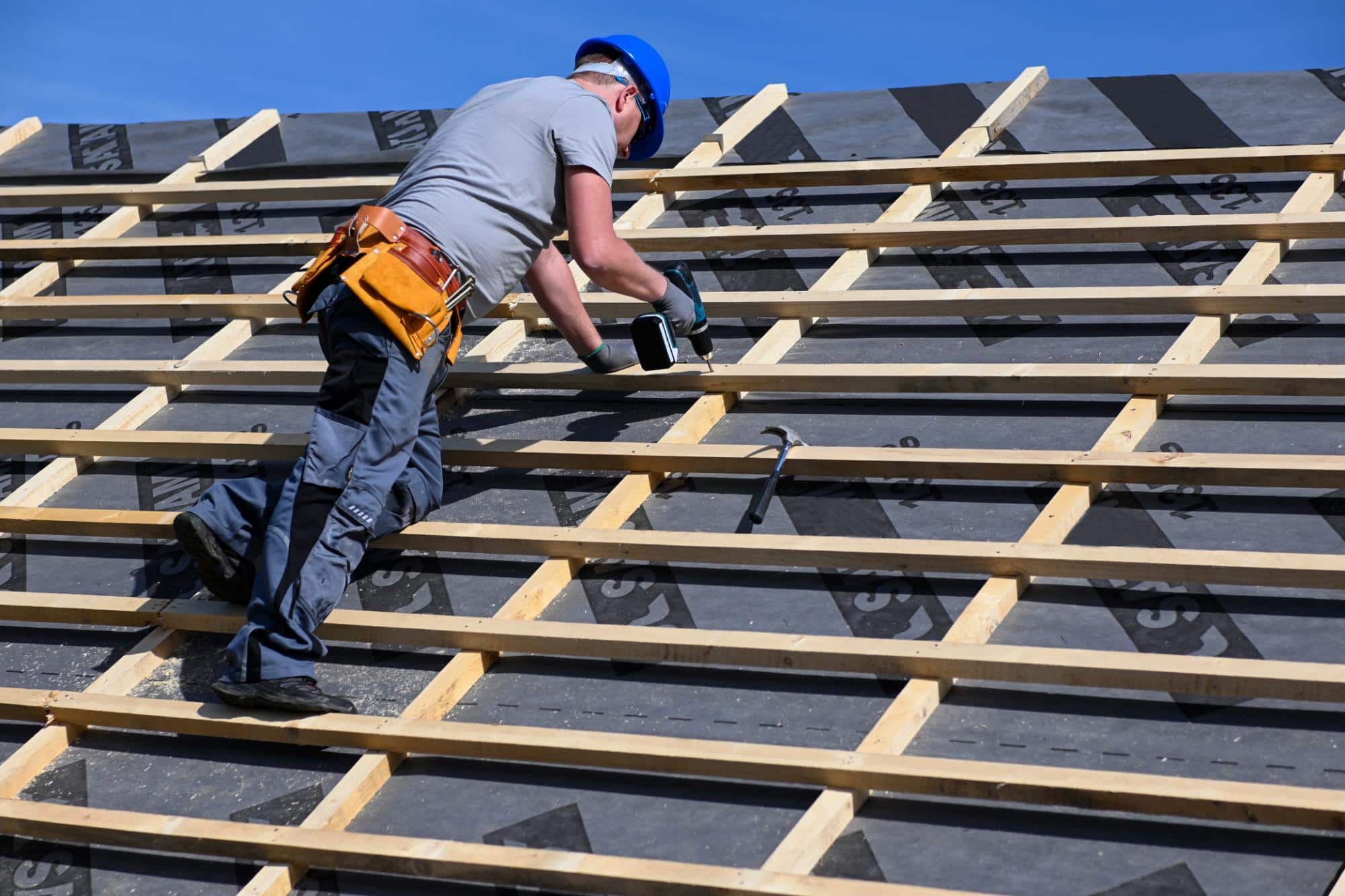 A construction worker wearing a blue hard hat and tool belt installs wooden battens on a sloped roof using a power drill, with clear blue sky in the background.