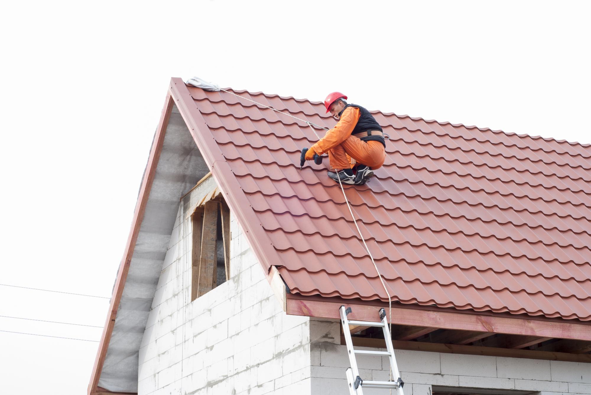 A worker in safety gear and a red helmet is fixing or inspecting a red metal roof while secured with a harness. A ladder is leaned against the side of the unfinished white brick building.