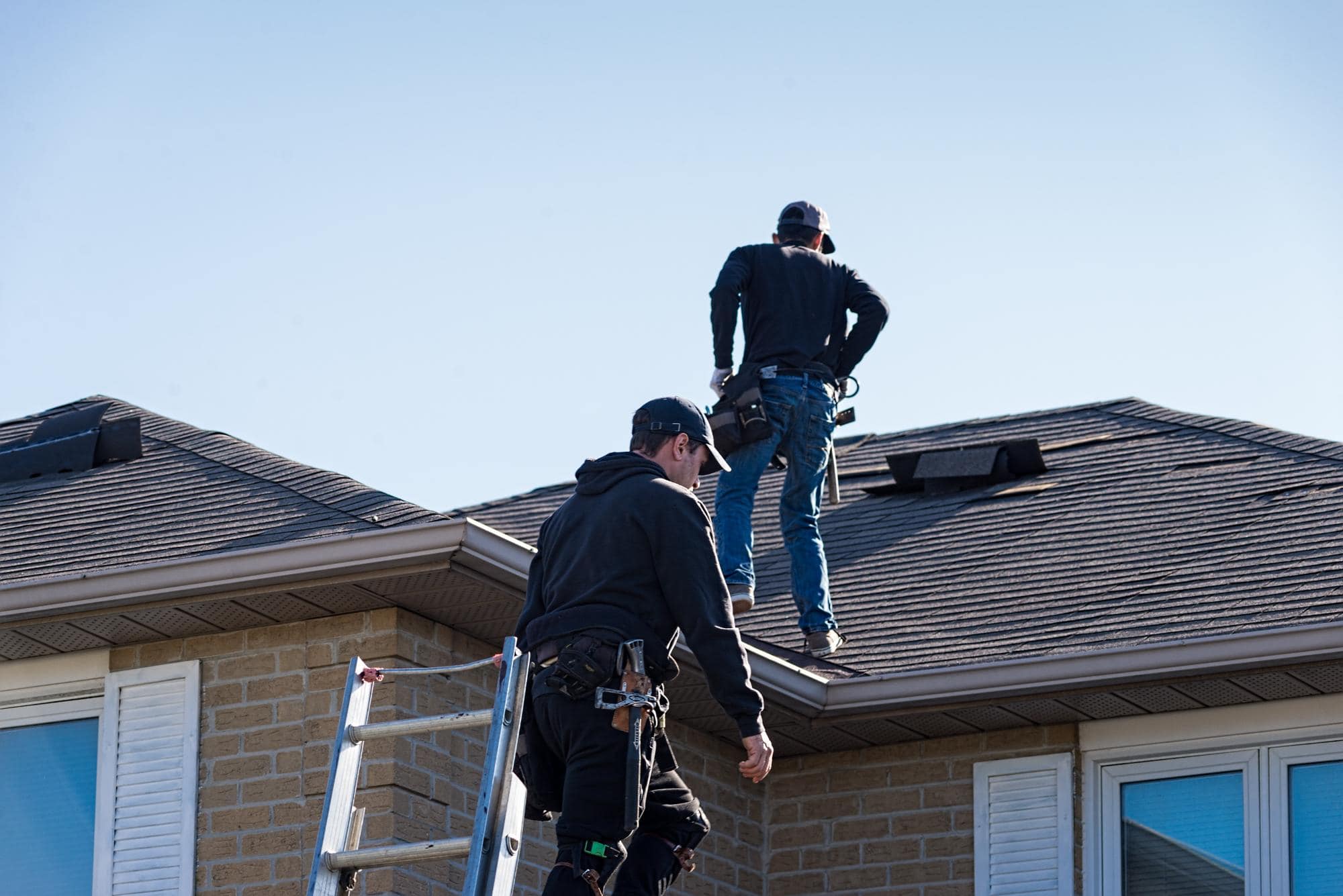 Two workers wearing safety gear are repairing or inspecting the roof of a house. One stands on a ladder, while the other is on the sloped roof, both with tools and equipment under a clear blue sky.