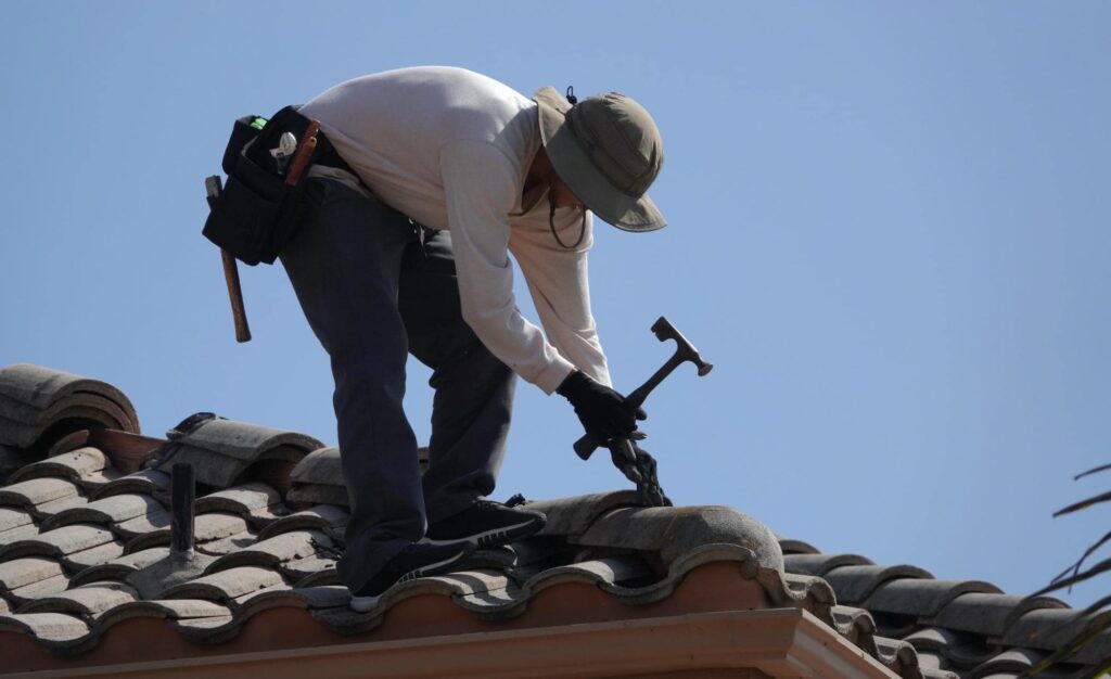 A worker wearing a hat, gloves, and a tool belt repairs roof tiles on a house under a clear blue sky, using a hammer.