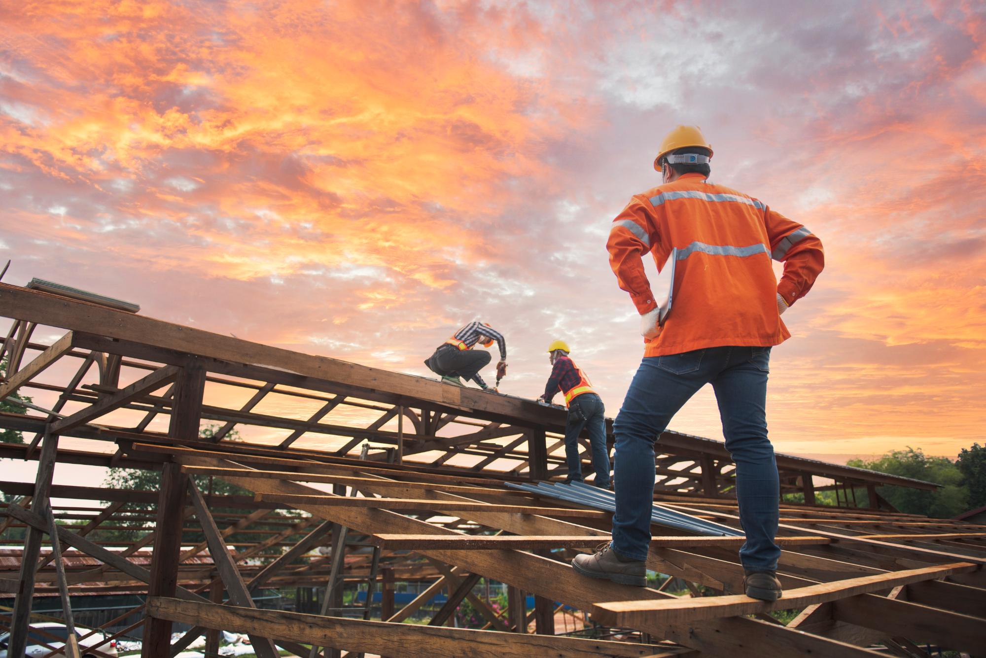 Construction workers wearing orange safety jackets and helmets work on the wooden framework of a building at sunset, with a dramatic sky of orange and pink clouds in the background.