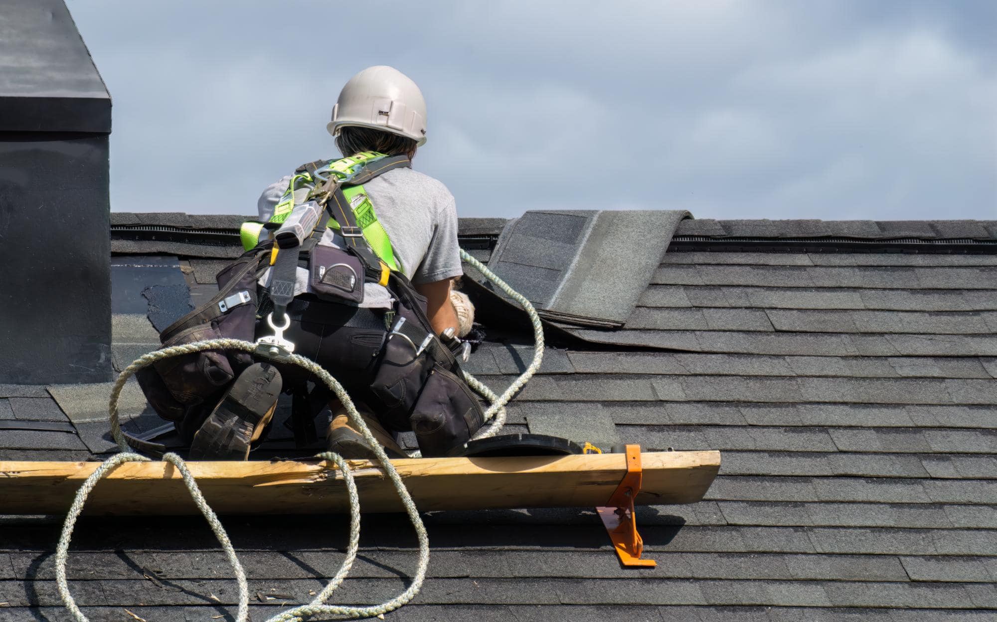 A construction worker with safety gear and a harness is kneeling on a roof, installing or repairing shingles. Safety ropes and wooden planks are used for support. The sky is cloudy in the background.