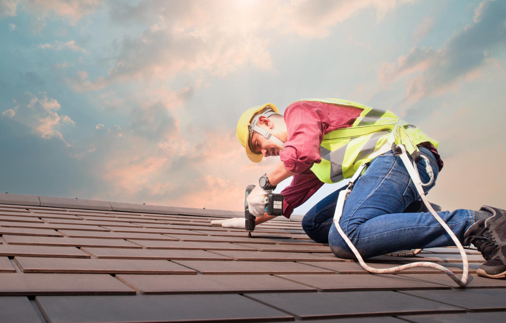 A construction worker wearing a safety helmet, vest, and harness kneels on a tiled roof, using a power drill. The sky in the background is partly cloudy with sunlight shining through.