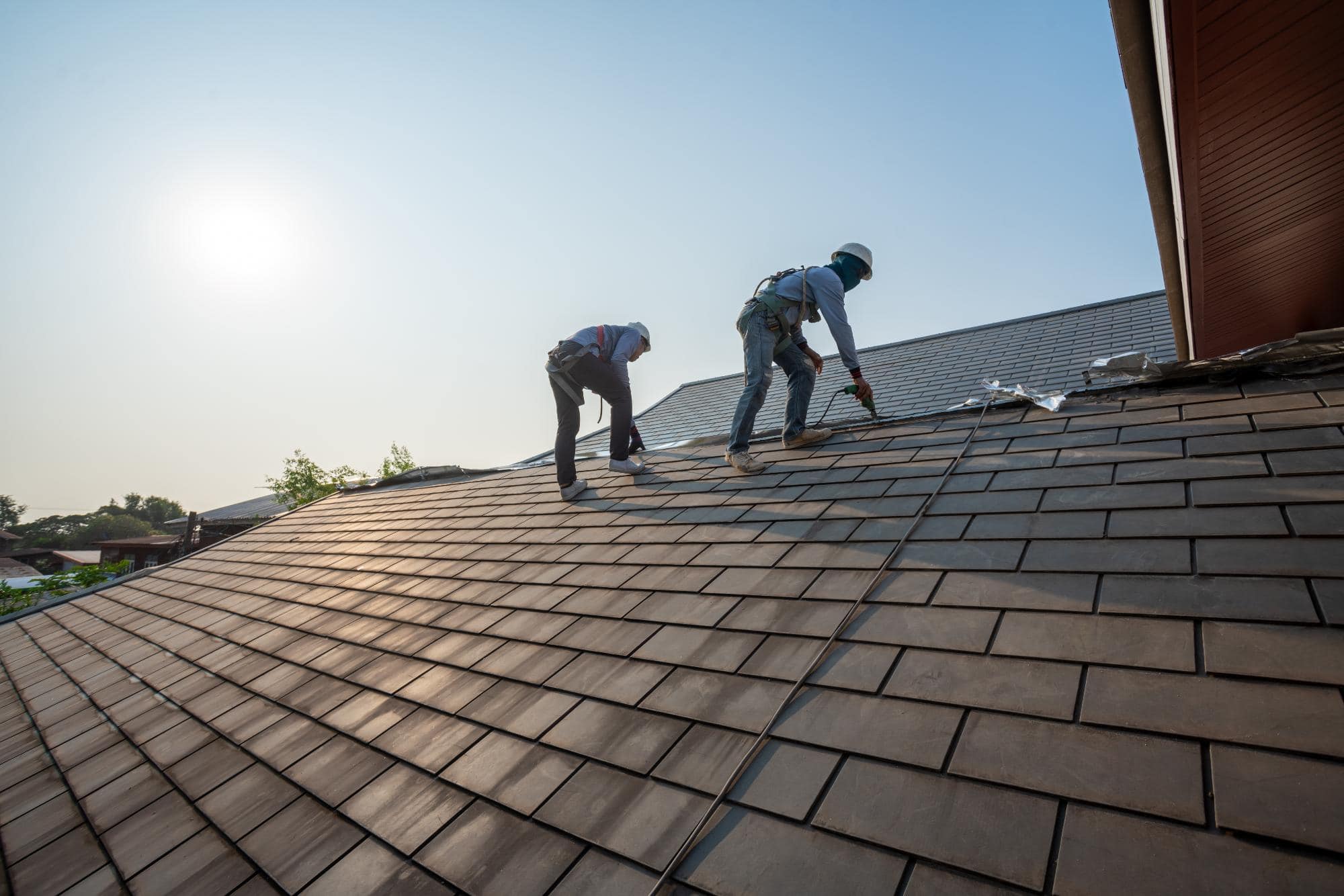 Two workers wearing safety harnesses are installing or repairing roof shingles on a sloped rooftop under a clear sky, with the sun shining in the background.