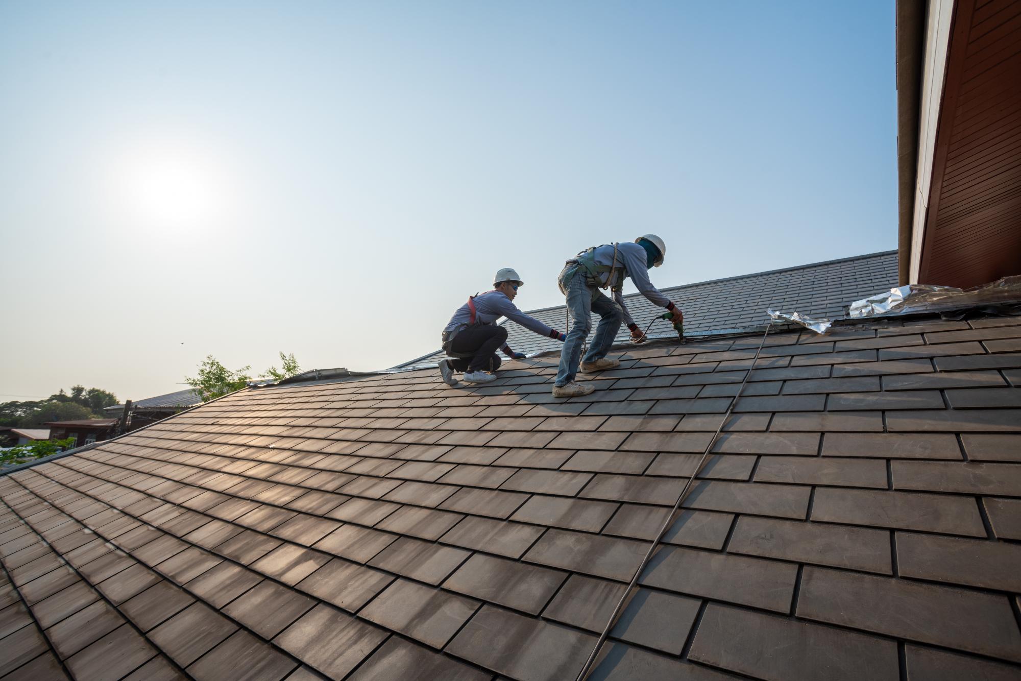 Two workers wearing safety gear are repairing or installing shingles on a sloped house roof under a clear sky, with the sun shining in the background.