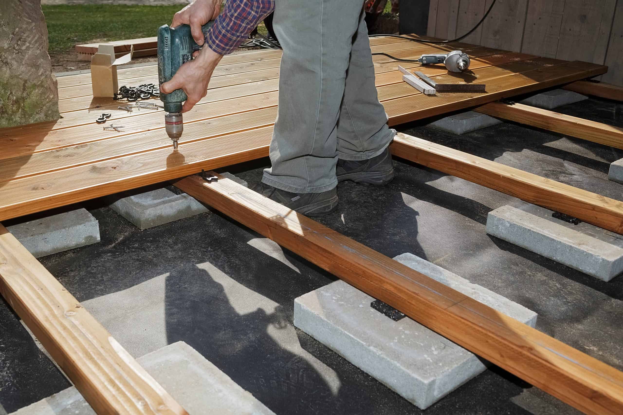 A person using a power drill to install wooden deck boards onto a frame supported by concrete blocks, with construction tools scattered nearby.