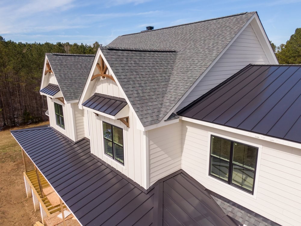 A modern farmhouse features a combination of gray asphalt shingles and dark metal roofing, with white siding and large black-framed windows, surrounded by trees.
