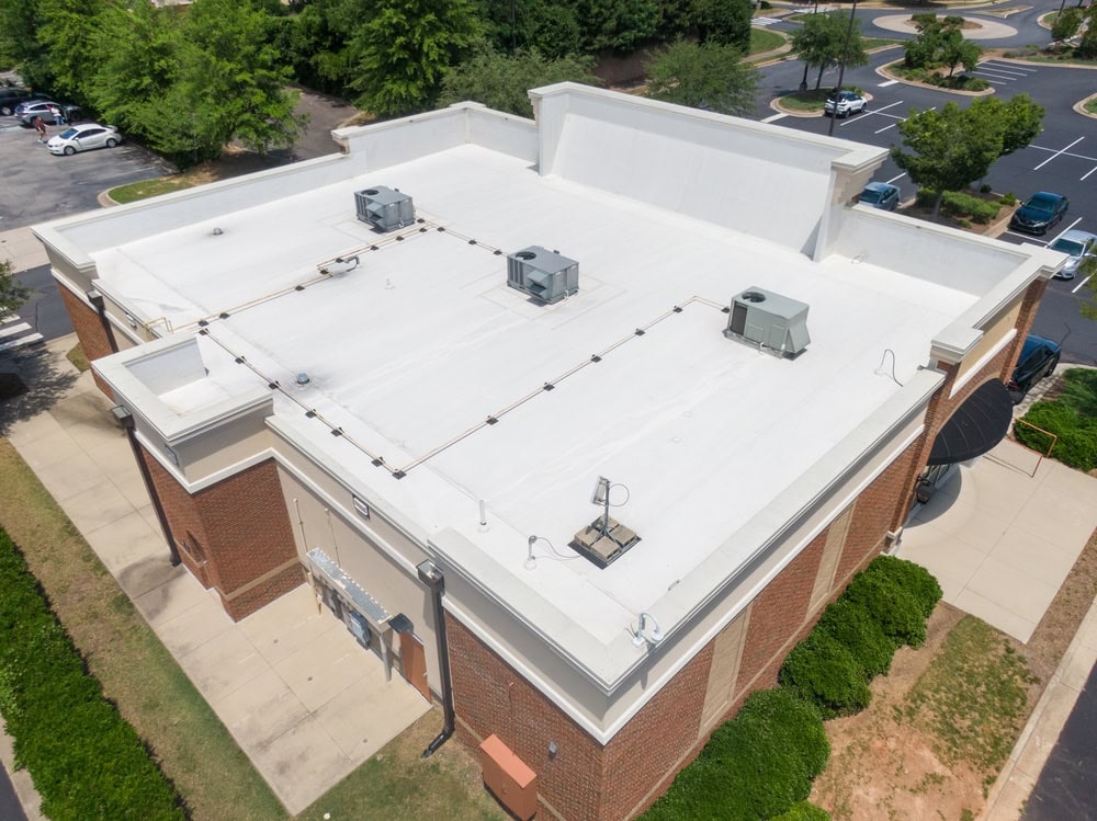 Aerial view of a flat-roofed brick building with three HVAC units and various pipes on the white roof, surrounded by sidewalks, landscaping, and nearby parking lots.