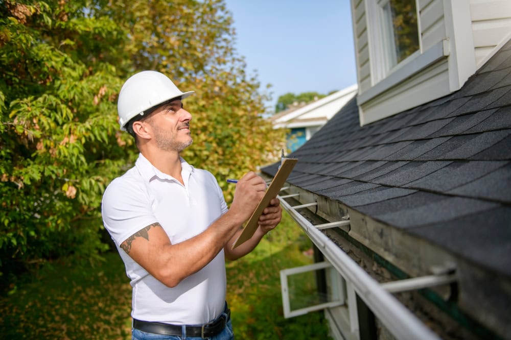 A man in a white hard hat and polo shirt smiles while inspecting a house's roof and gutters, holding a clipboard and pen, with trees and greenery in the background.