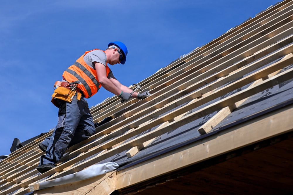 A construction worker in a blue hard hat and orange safety vest installs wooden beams on a sloped roof under a clear blue sky.