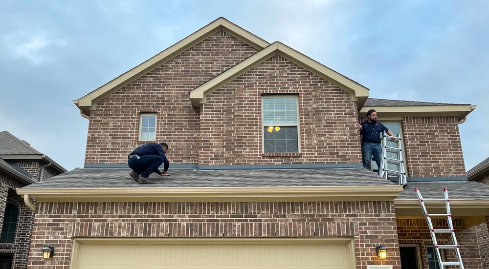 Two people are inspecting the roof of a two-story brick house; one person is crouched on the roof while the other stands and holds a ladder near the edge. The sky is mostly cloudy.
